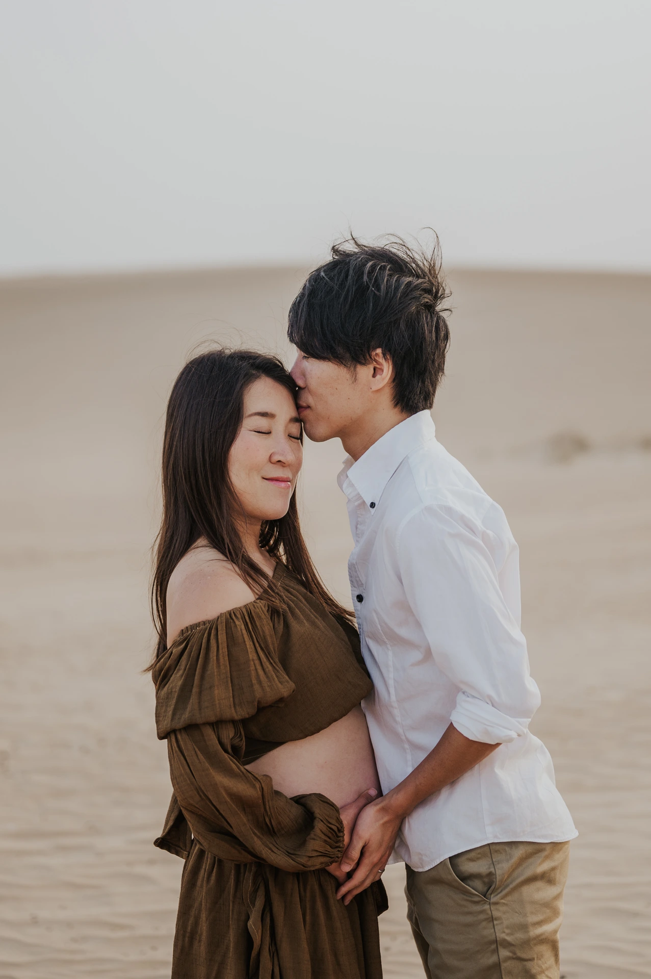 An expecting couple poses on Dubai dunes during a maternity photoshoot, with the partner kissing her forehead while both hold the baby bump.