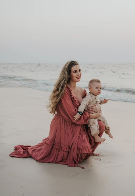 A mother kneels on Dubai beach holding her toddler during a maternity and family photoshoot, with shoreline waves and soft evening light behind them.