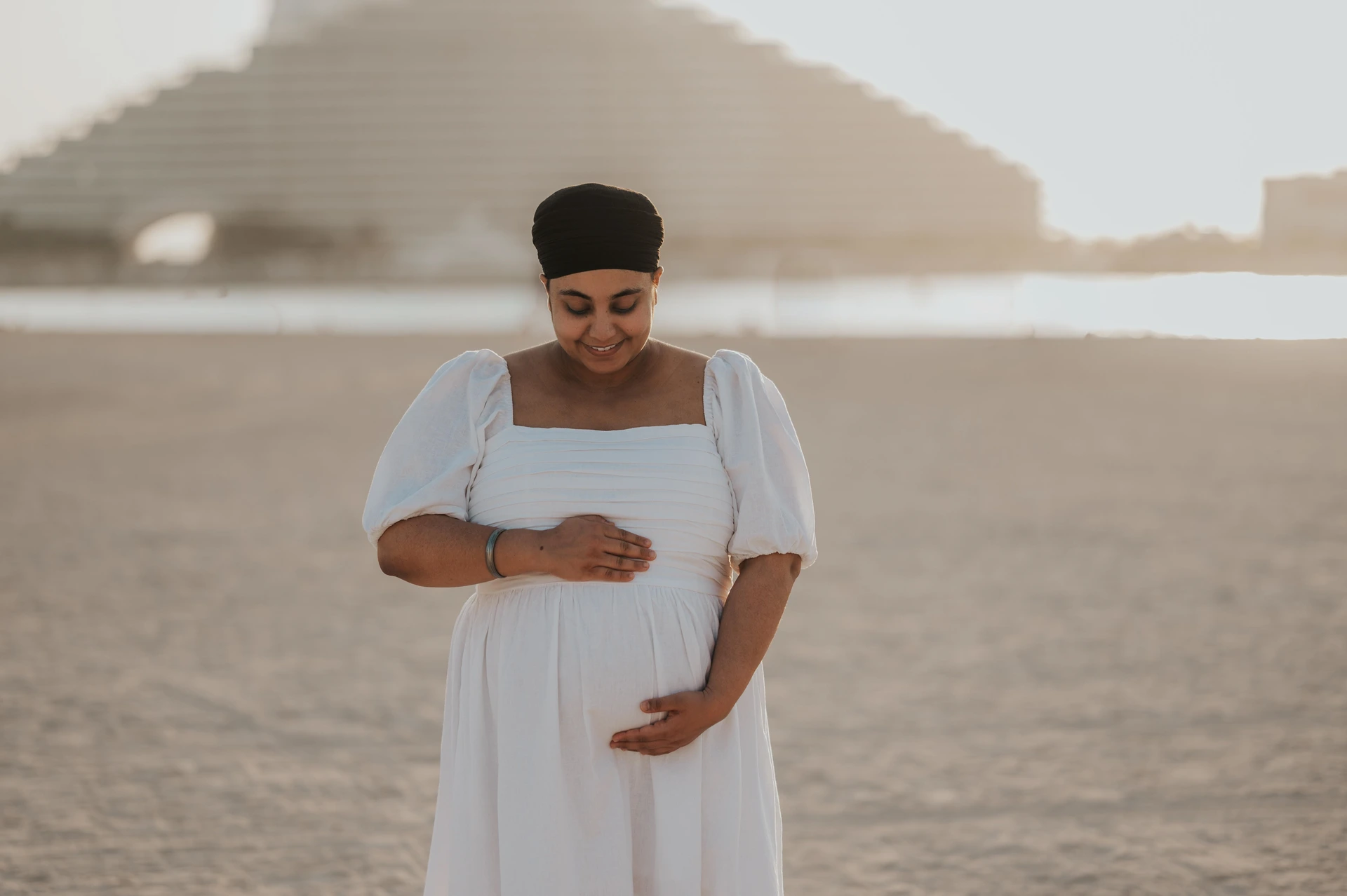 A pregnant woman stands on Dubai beach with Burj Al Arab behind her during a maternity photoshoot, captured in warm low-angle light.