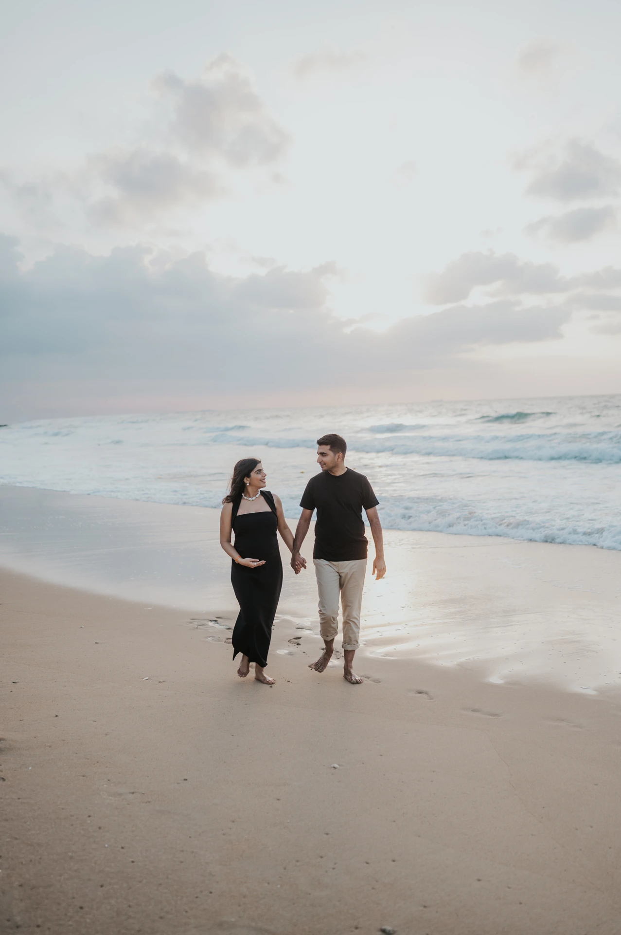 An expecting couple holds hands and walks along the Dubai shoreline in a maternity photoshoot, showing a balanced frame for sunrise and sunset mood.