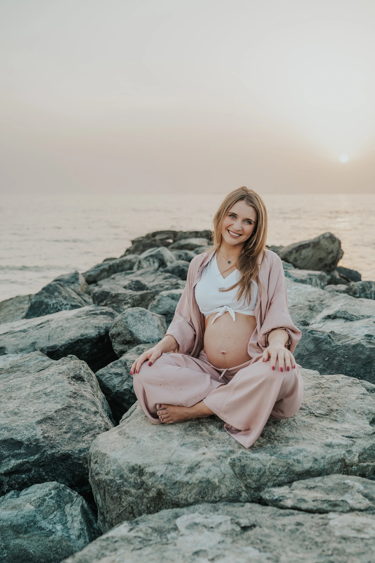 A pregnant woman sits on shoreline rocks in Dubai during a maternity photoshoot, creating a relaxed solo portrait with soft sea and sky backdrop.