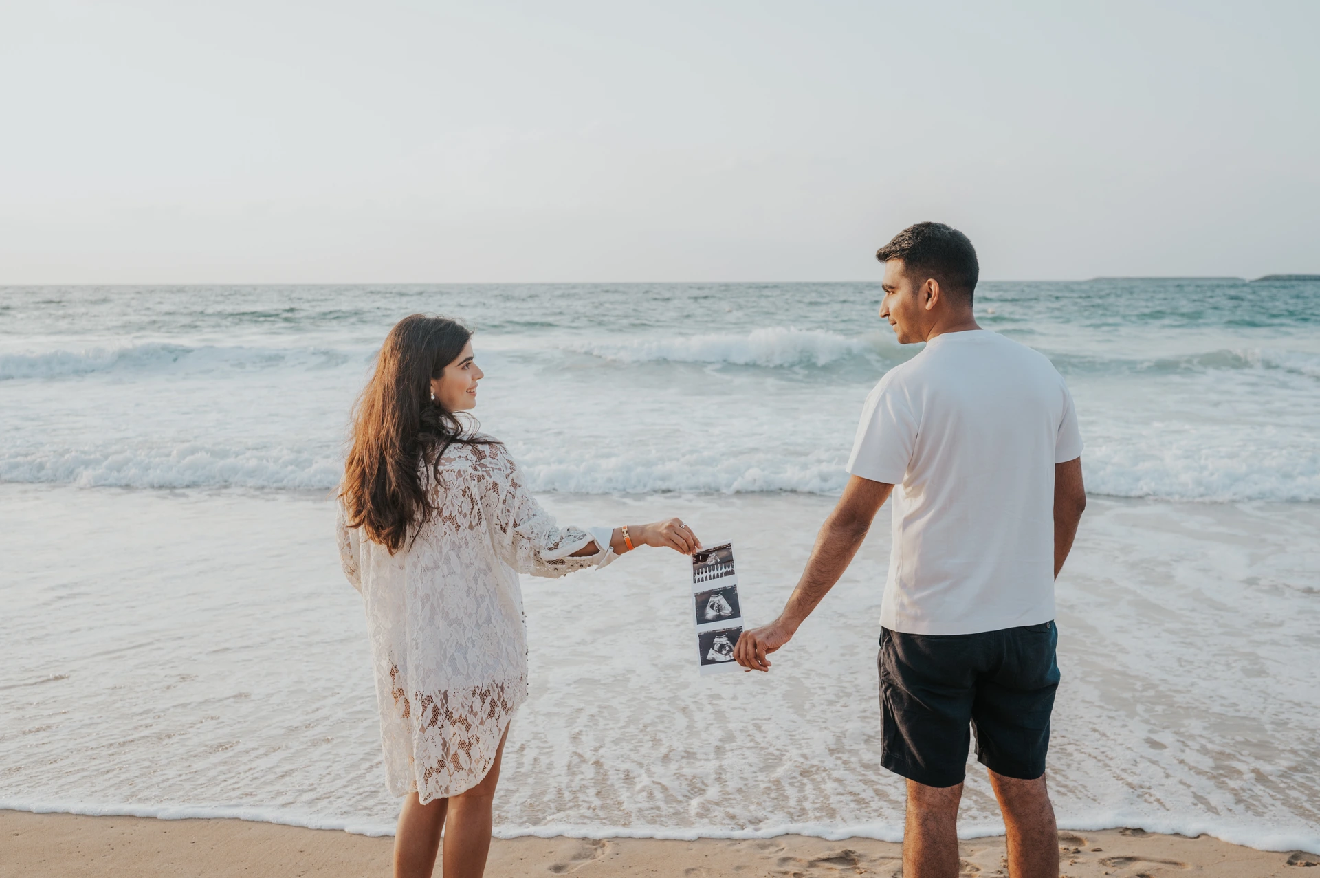 An expecting couple walks at the Dubai shoreline while holding ultrasound prints during a maternity photoshoot, with soft light and gentle waves behind them.