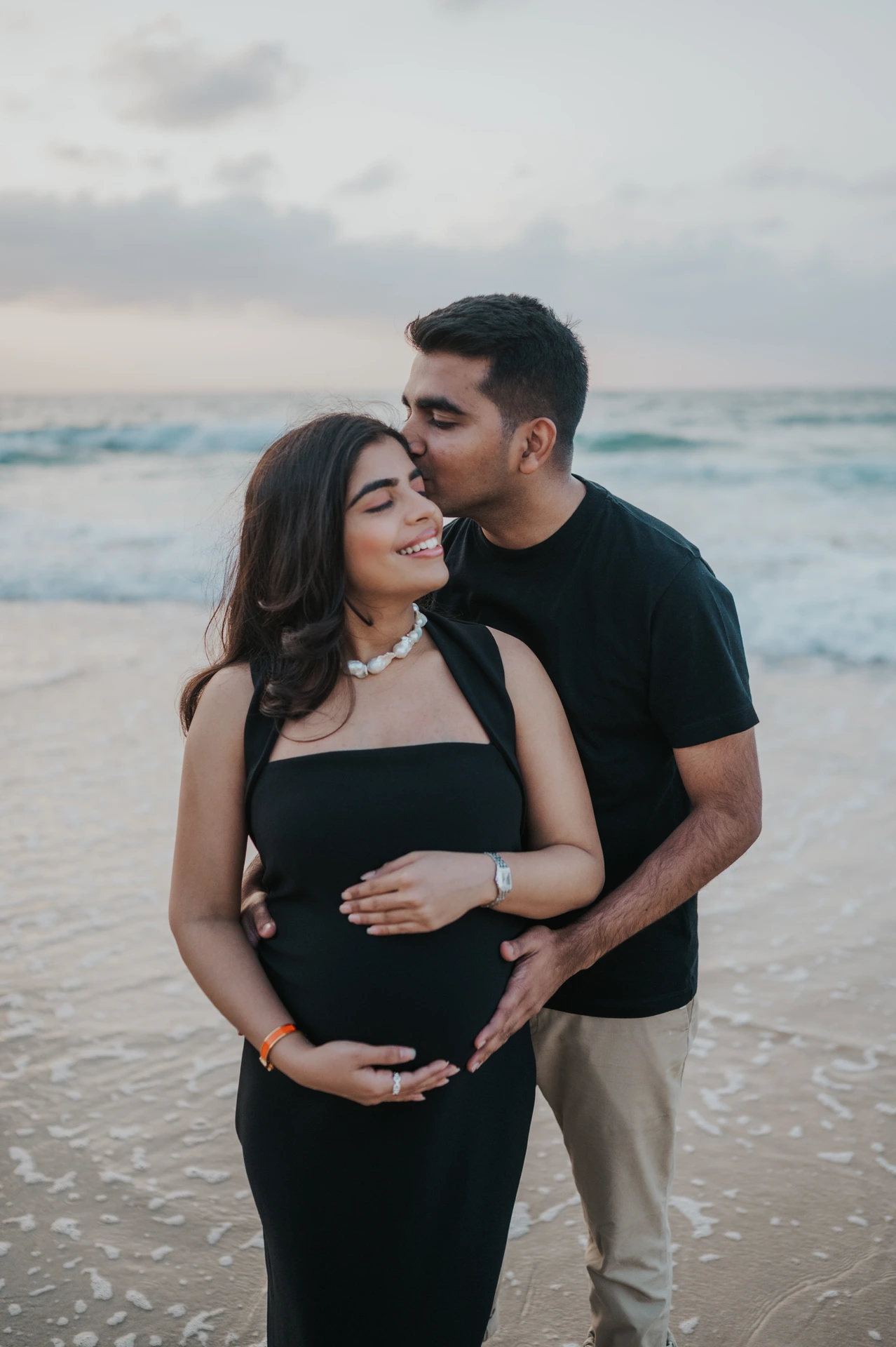 A couple stands close at the Dubai shoreline during a maternity shoot, with the mother in a fitted black dress that keeps shape against sea wind.