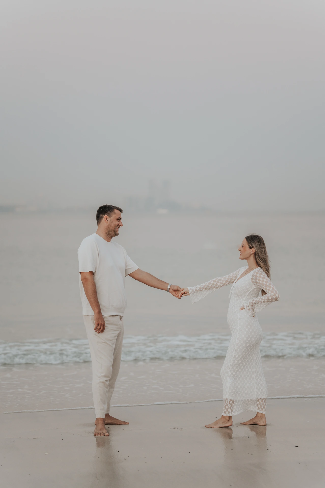 A pregnant woman in a white lace dress holds hands with her partner on Dubai beach at sunset.