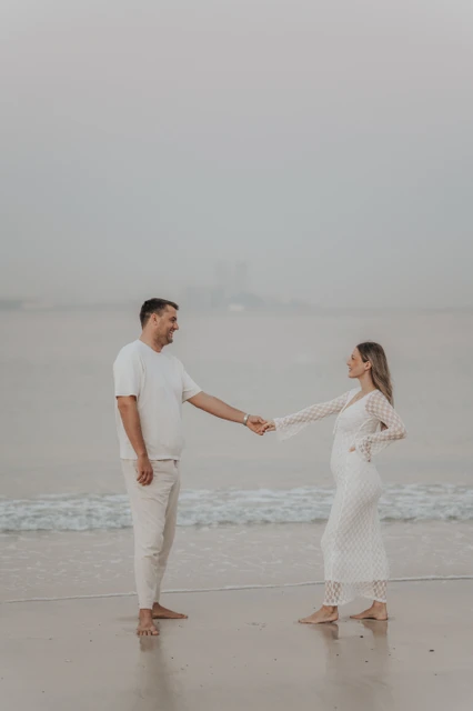 Dubai maternity outfit inspiration - beach session A pregnant woman in a white lace dress holds hands with her partner on Dubai beach at sunset.