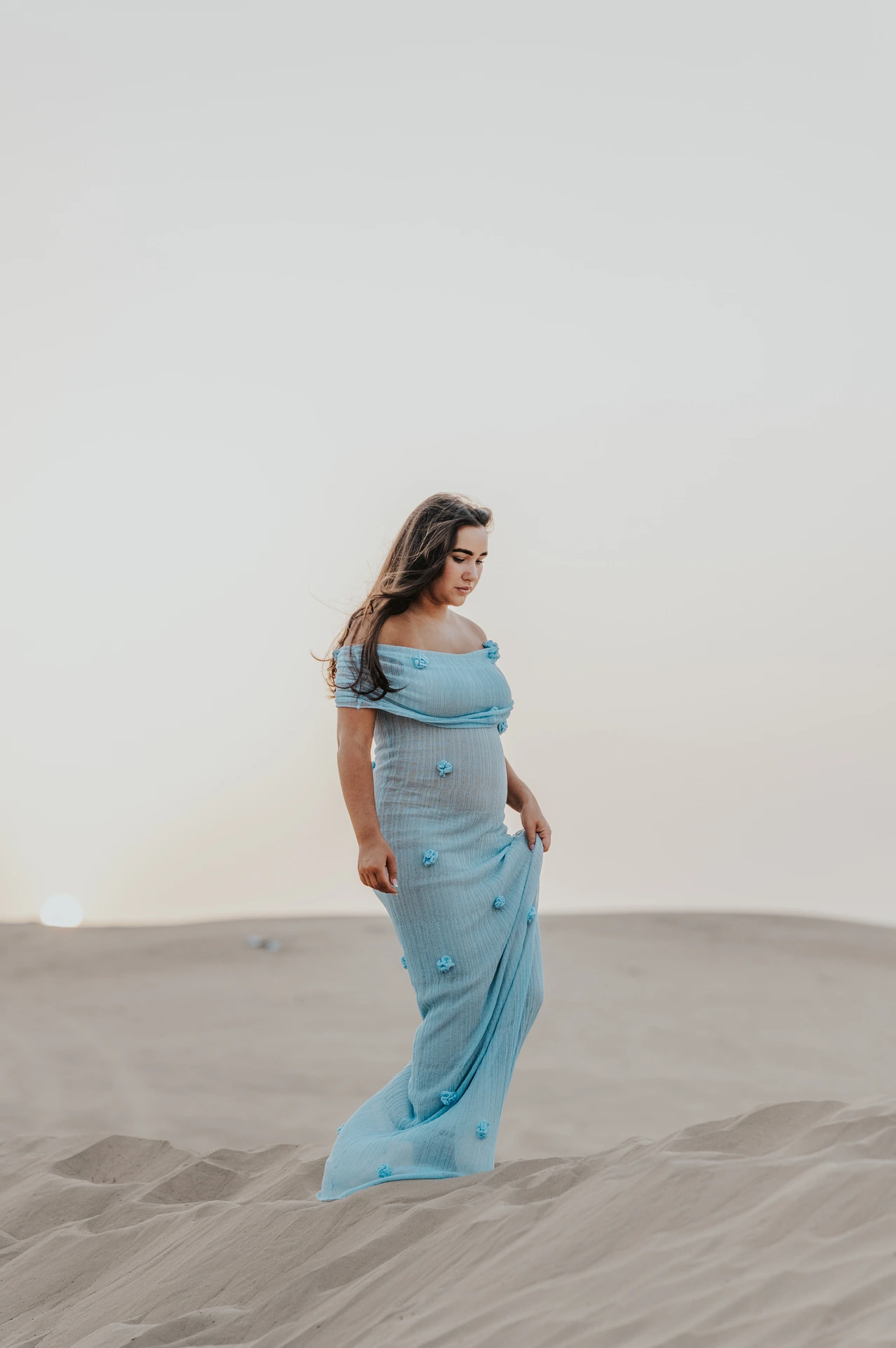 A pregnant woman walks on Dubai desert dunes during a maternity shoot, wearing a flowing blue gown that adds movement and texture in open sand light.