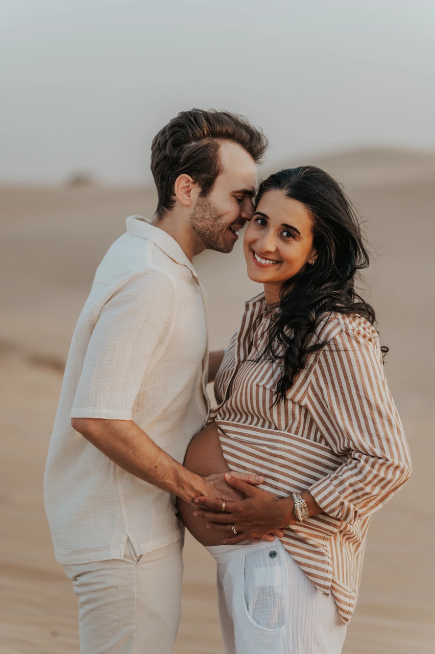 Babymoon Photoshoot Dubai - desert forehead kiss An expecting mother smiles at the camera while her partner kisses her forehead on Dubai dunes during a babymoon photoshoot, both hands resting on her baby bump.