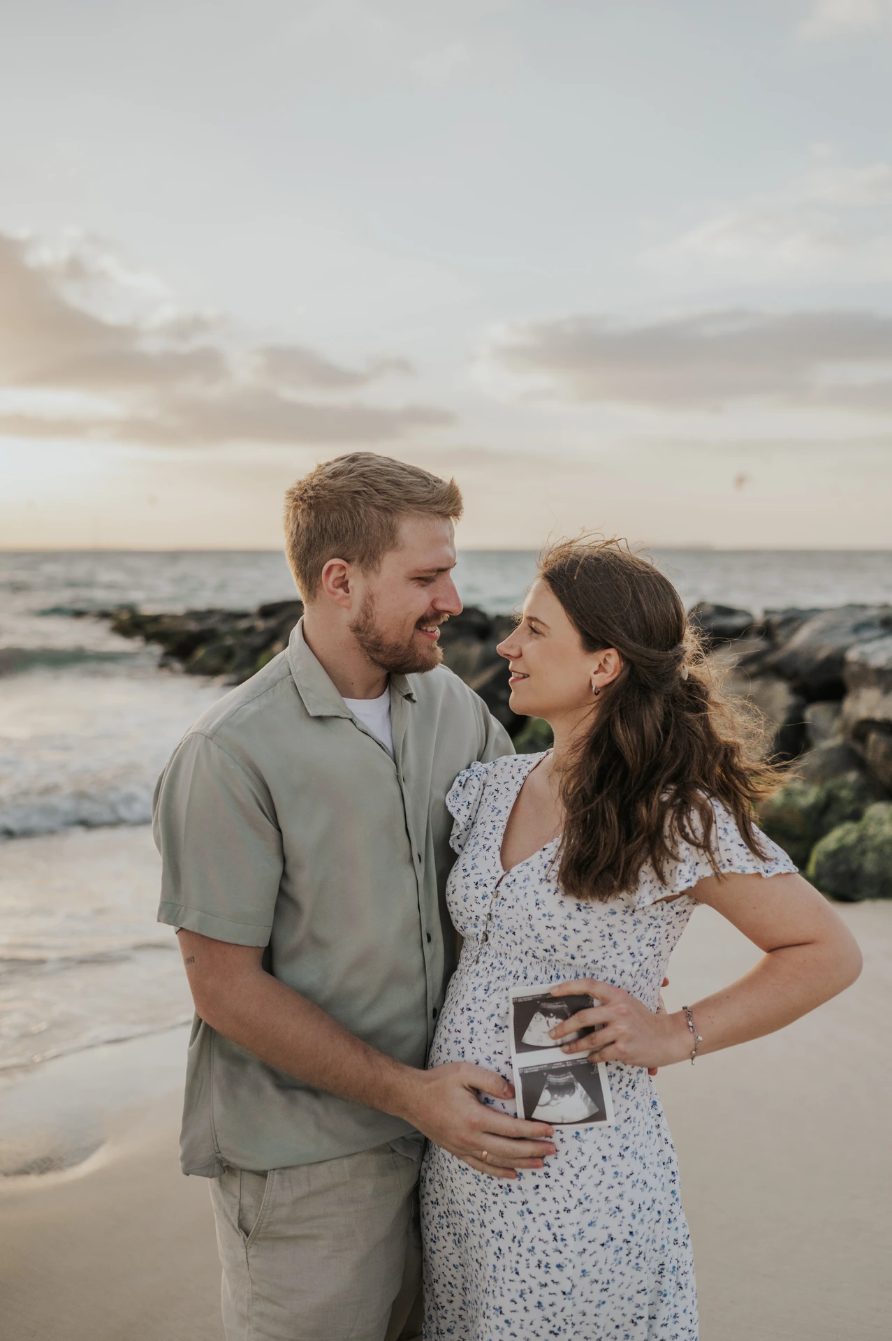 An expecting couple embraces on Dubai beach during a babymoon photoshoot, with ultrasound prints held near the baby bump and rocky shoreline behind them.
