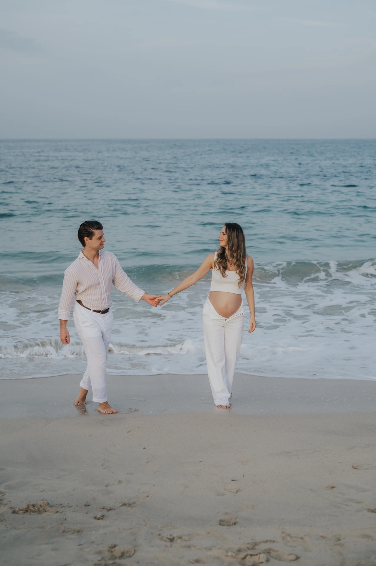 An expecting couple walks hand in hand at the Dubai waterline during a babymoon photoshoot, with turquoise waves behind them and coordinated white outfits.