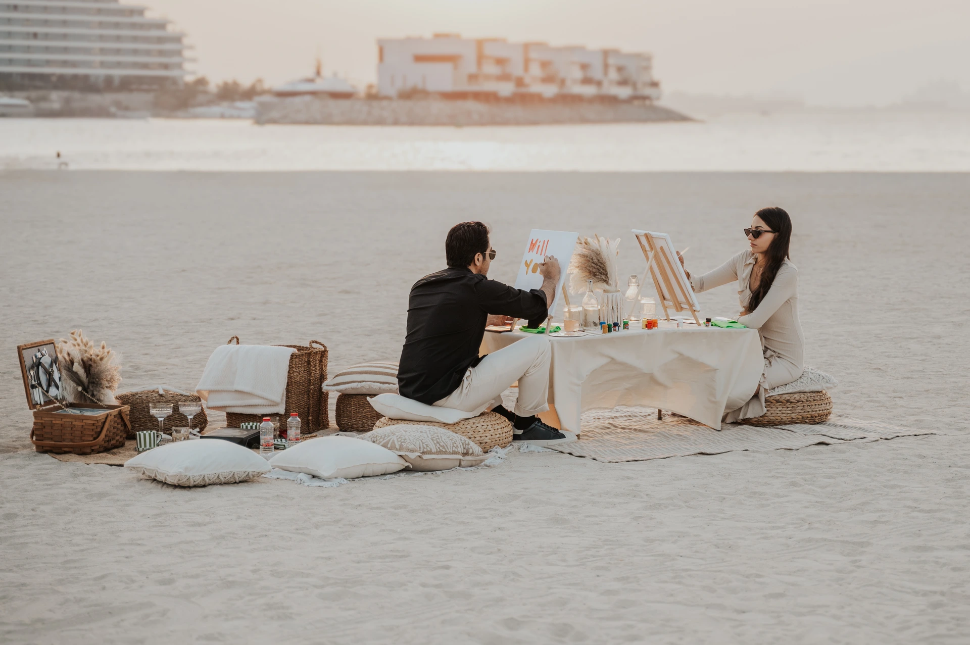 Dubai Photohoot - beach picnic proposal setup A couple sits at a styled beach picnic setup in Dubai during a photoshoot, creating a romantic pre-proposal atmosphere by the water.