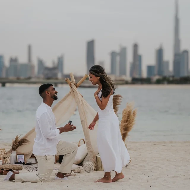 A man presents an open ring box beside a styled beach picnic with the Dubai skyline behind their proposal portrait.