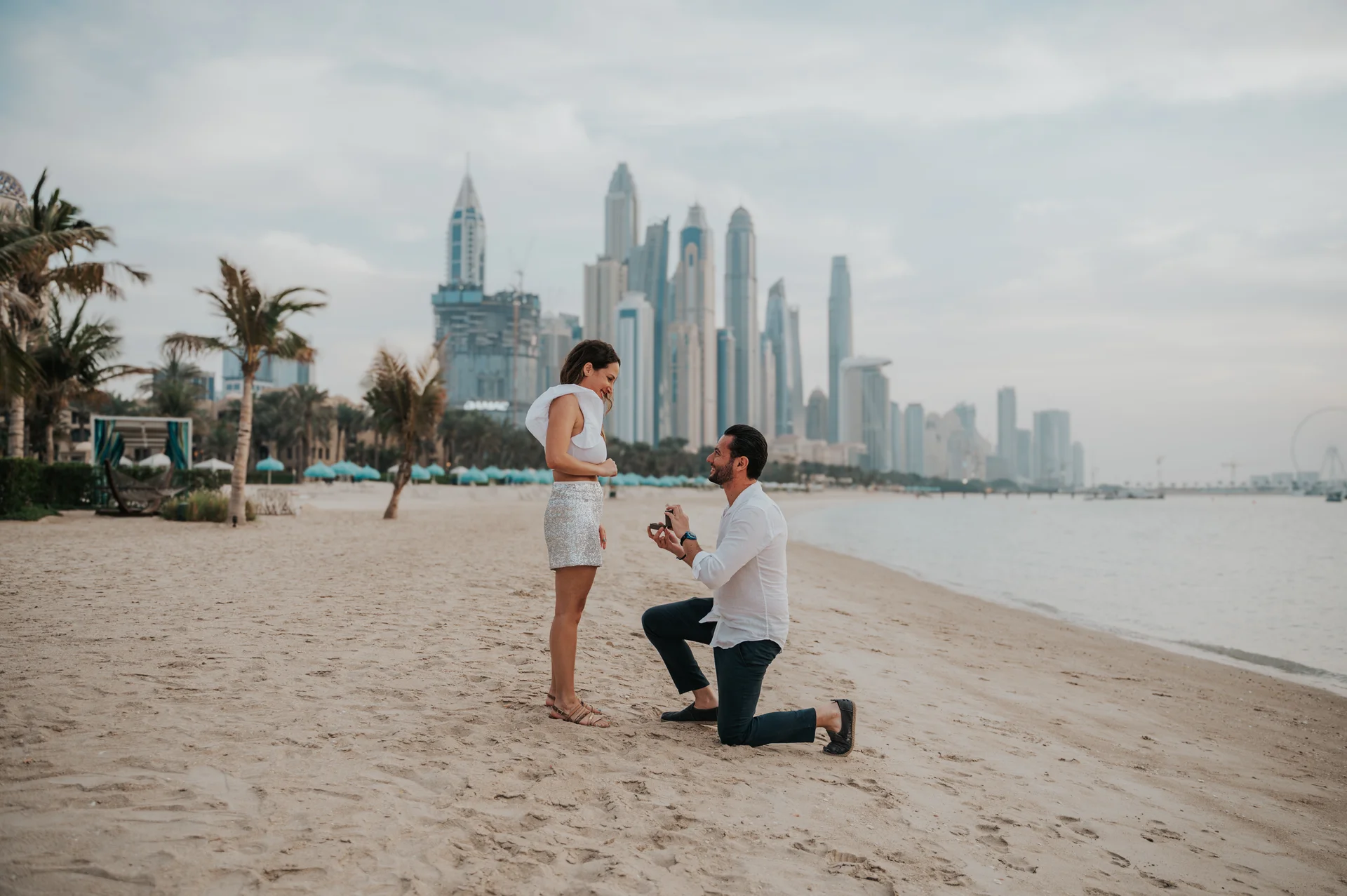 Proposing Before the Dubai Marina Skyline A man kneels with an open ring box on a beach before the Dubai Marina skyline during a surprise proposal photoshoot.