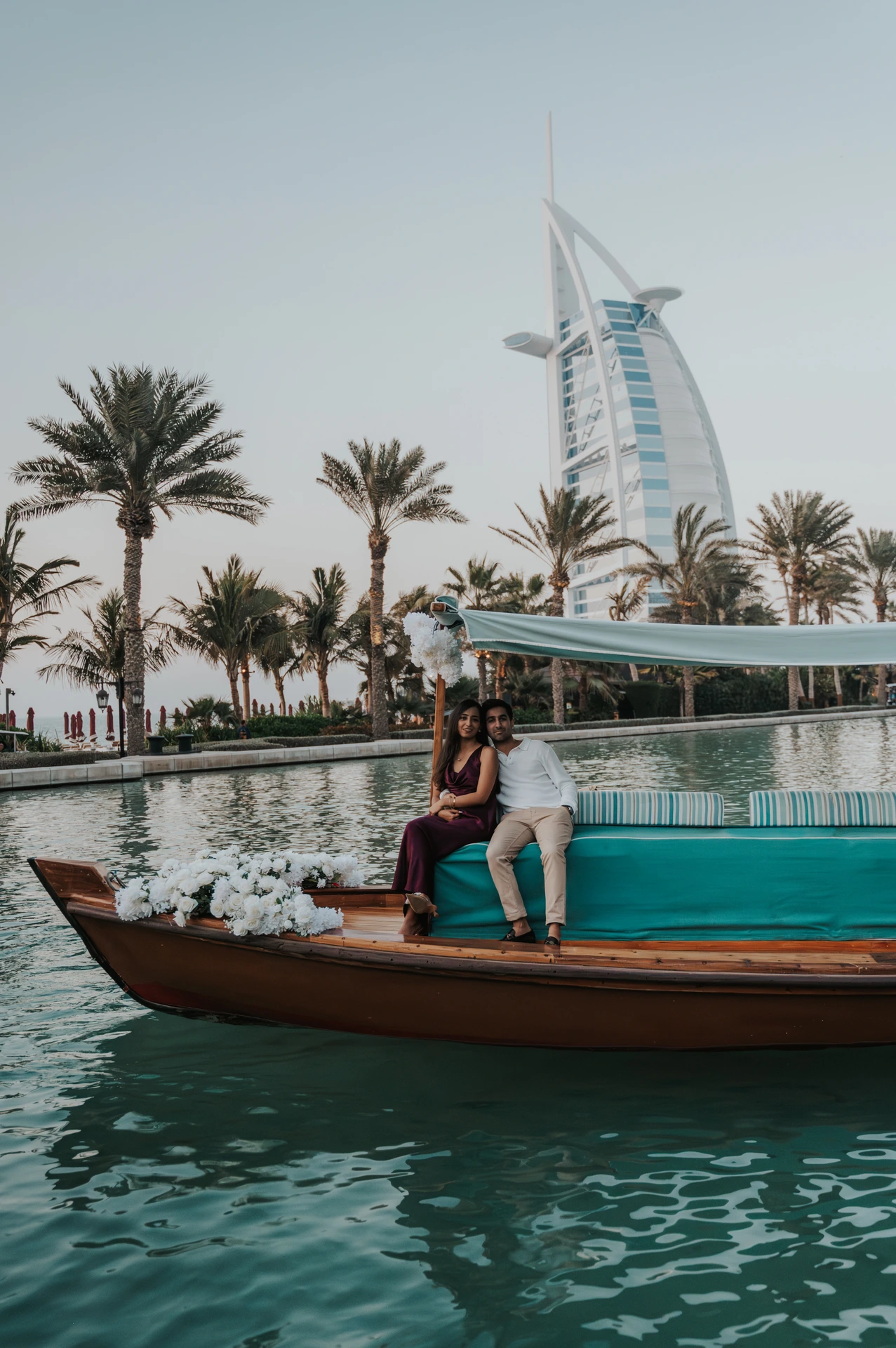 A couple sits together on a decorated abra near Shimmers in Dubai during a proposal-themed portrait with palms and water behind them.