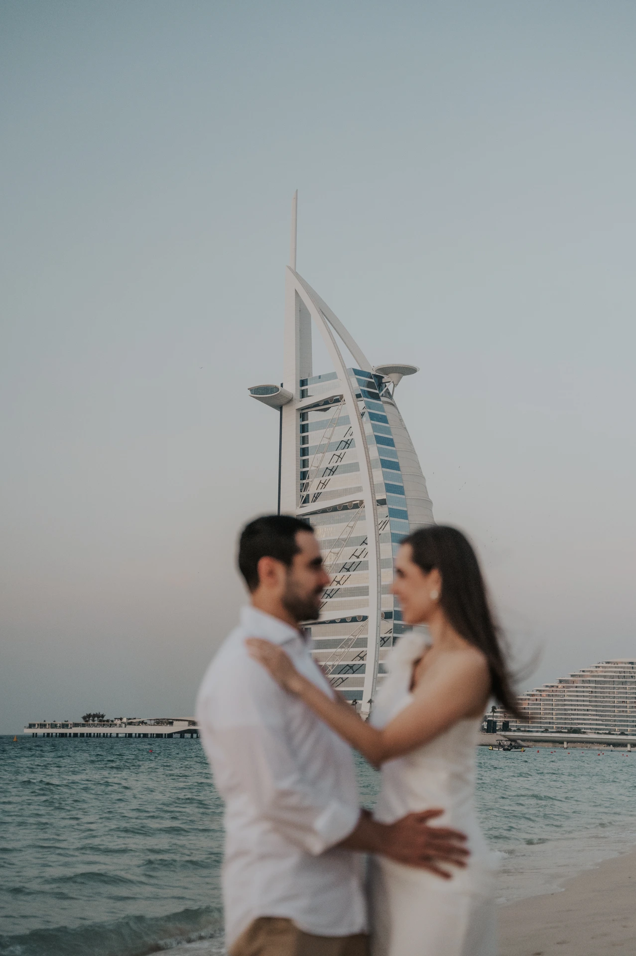 A couple stands close together on Dubai beach near Shimmers with Burj Al Arab in soft focus during their proposal session.