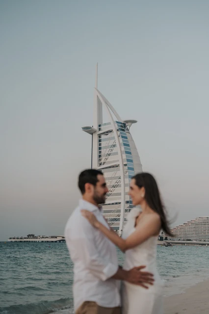 A couple stands close together on Dubai beach near Shimmers with Burj Al Arab in soft focus during their proposal session.