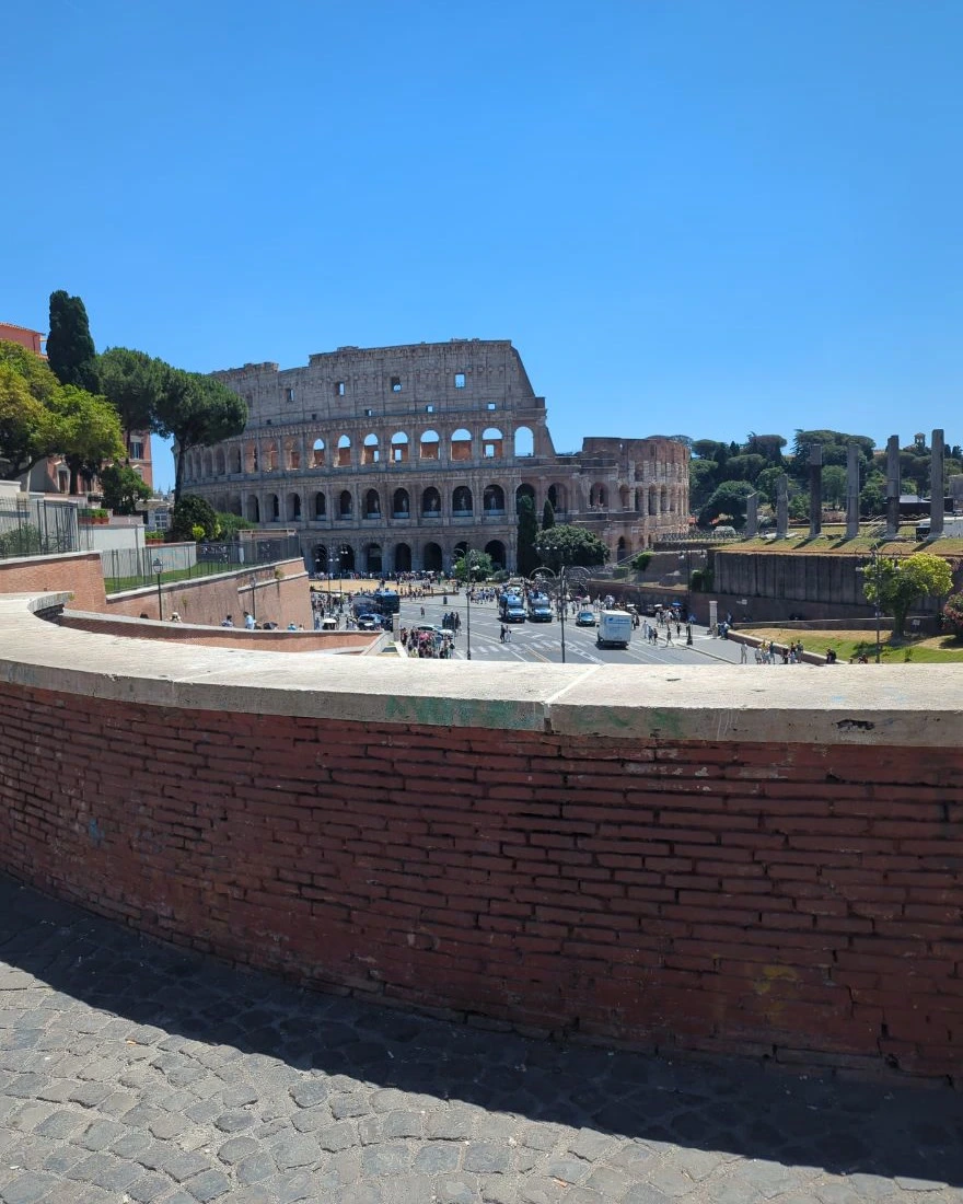 The Colosseum sits beyond a curved brick wall and cobbled foreground, showing a spacious elevated angle for a Rome photoshoot.