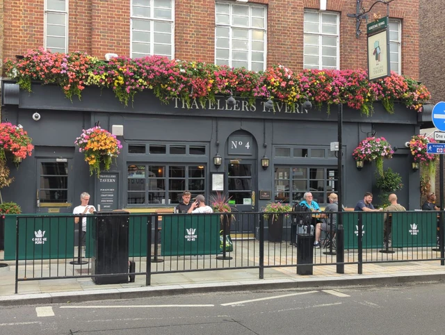 Dark grey Travellers Tavern frontage with number four above the door, bright hanging baskets and outdoor diners
