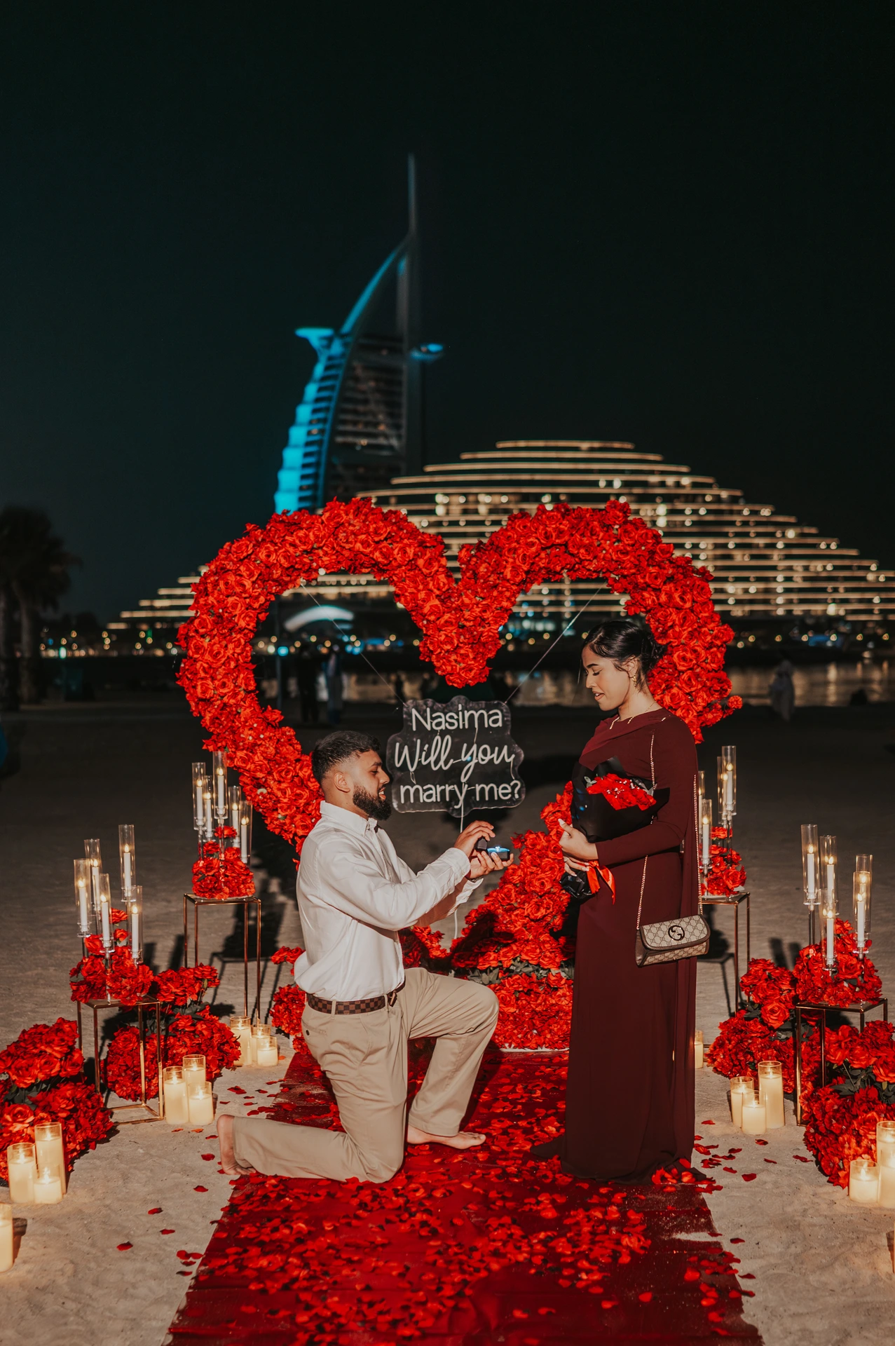 At night on Dubai beach, a man kneels in front of a red floral heart setup while proposing, with Burj Al Arab glowing behind the scene.