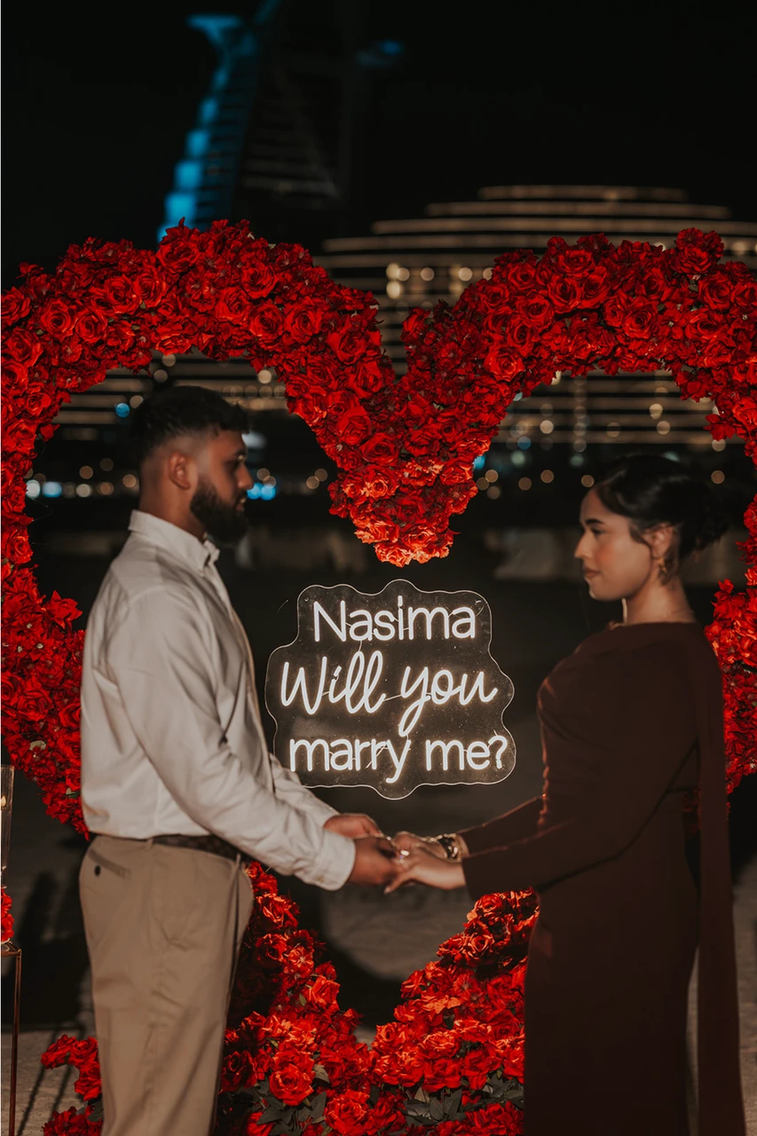 Heart Proposal Setup Dubai - hand-holding portrait inside red heart A couple holds hands inside a red floral heart setup on Dubai beach at night, standing in front of the lit proposal sign.