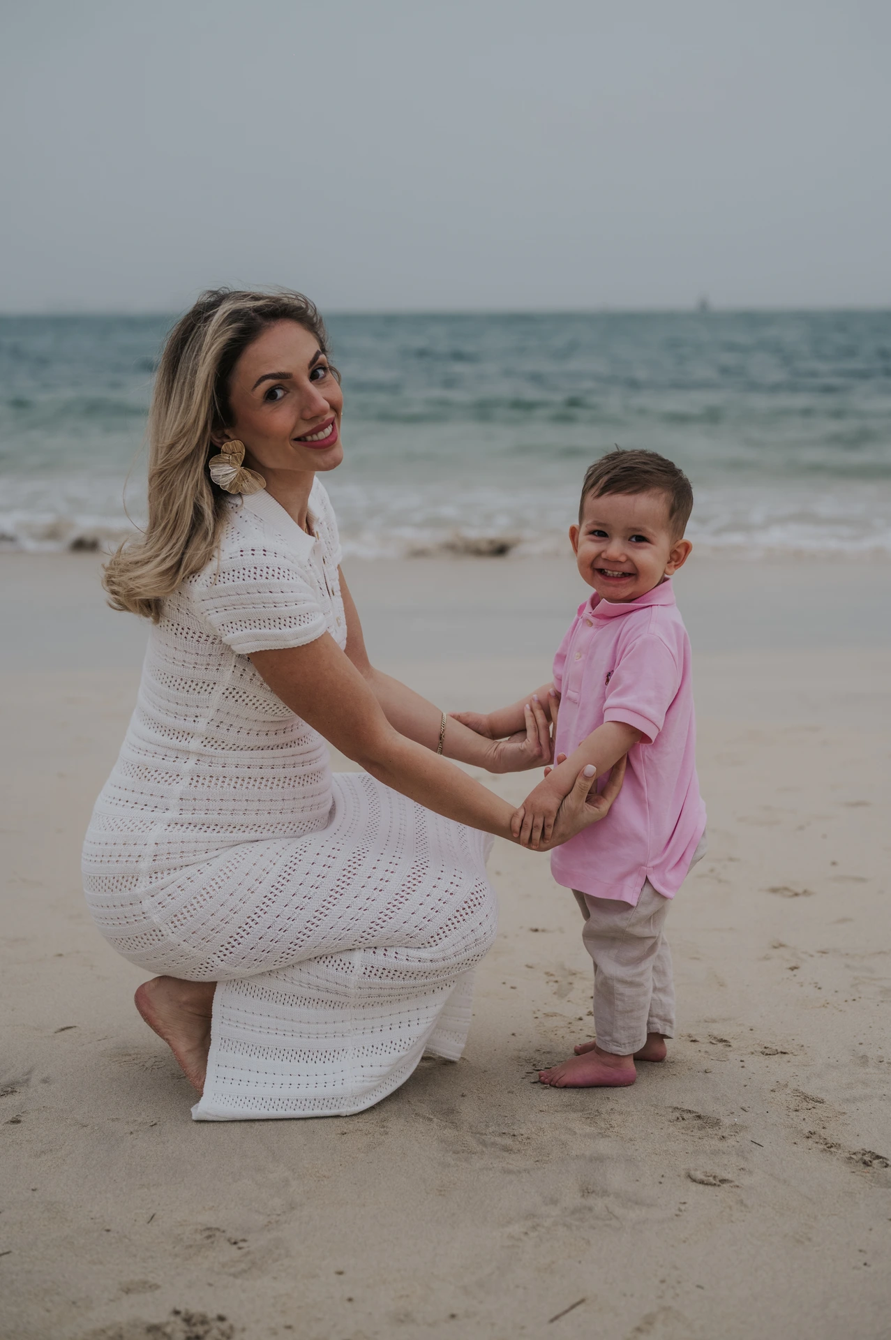 A mother in a white dress holds her little boy’s hands on Dubai beach during a warm family beach photoshoot.