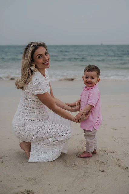 A mother in a white dress holds her little boy’s hands on Dubai beach during a warm family beach photoshoot.