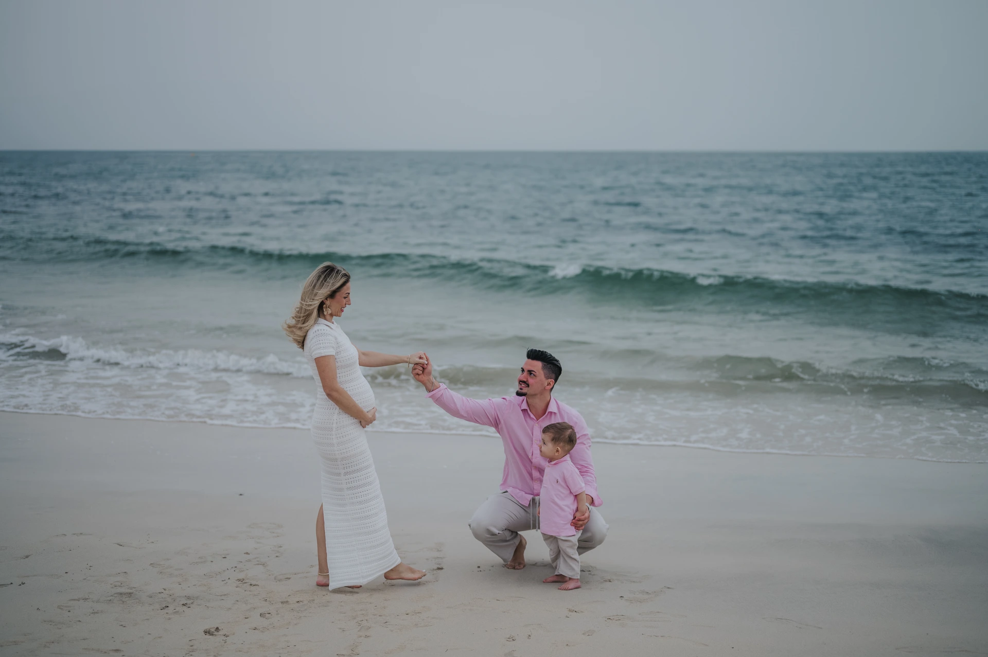 Parents kneel and reach toward their toddler by the waves on Dubai beach during a candid family beach photoshoot.