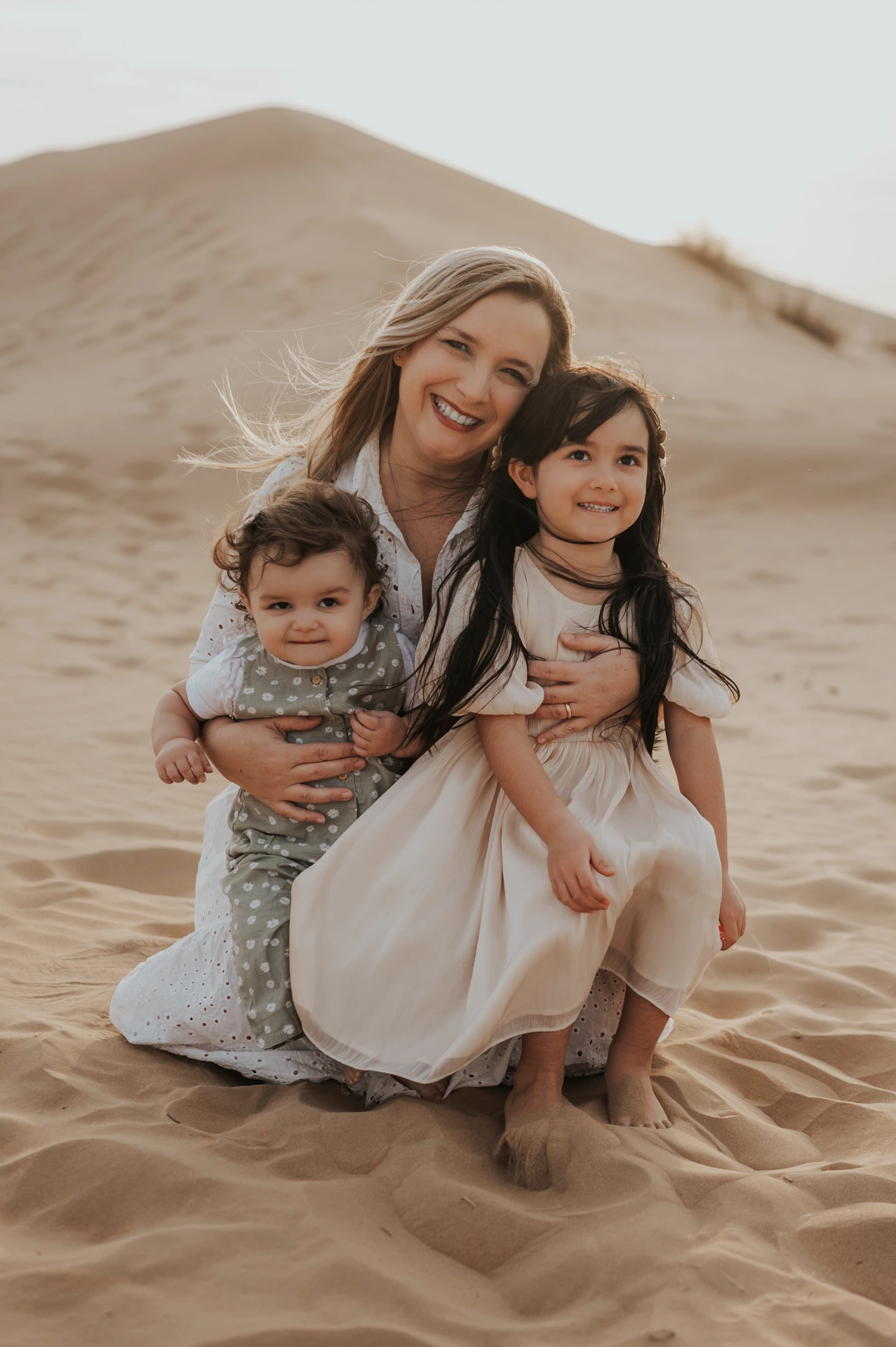 Mother hugging two young daughters on sandy dunes during a Dubai family photoshoot in warm evening light.