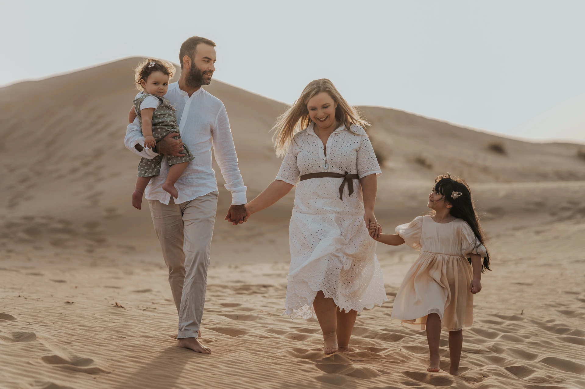 Parents and two children walk hand in hand across sunlit Dubai dunes during a relaxed family photo session.