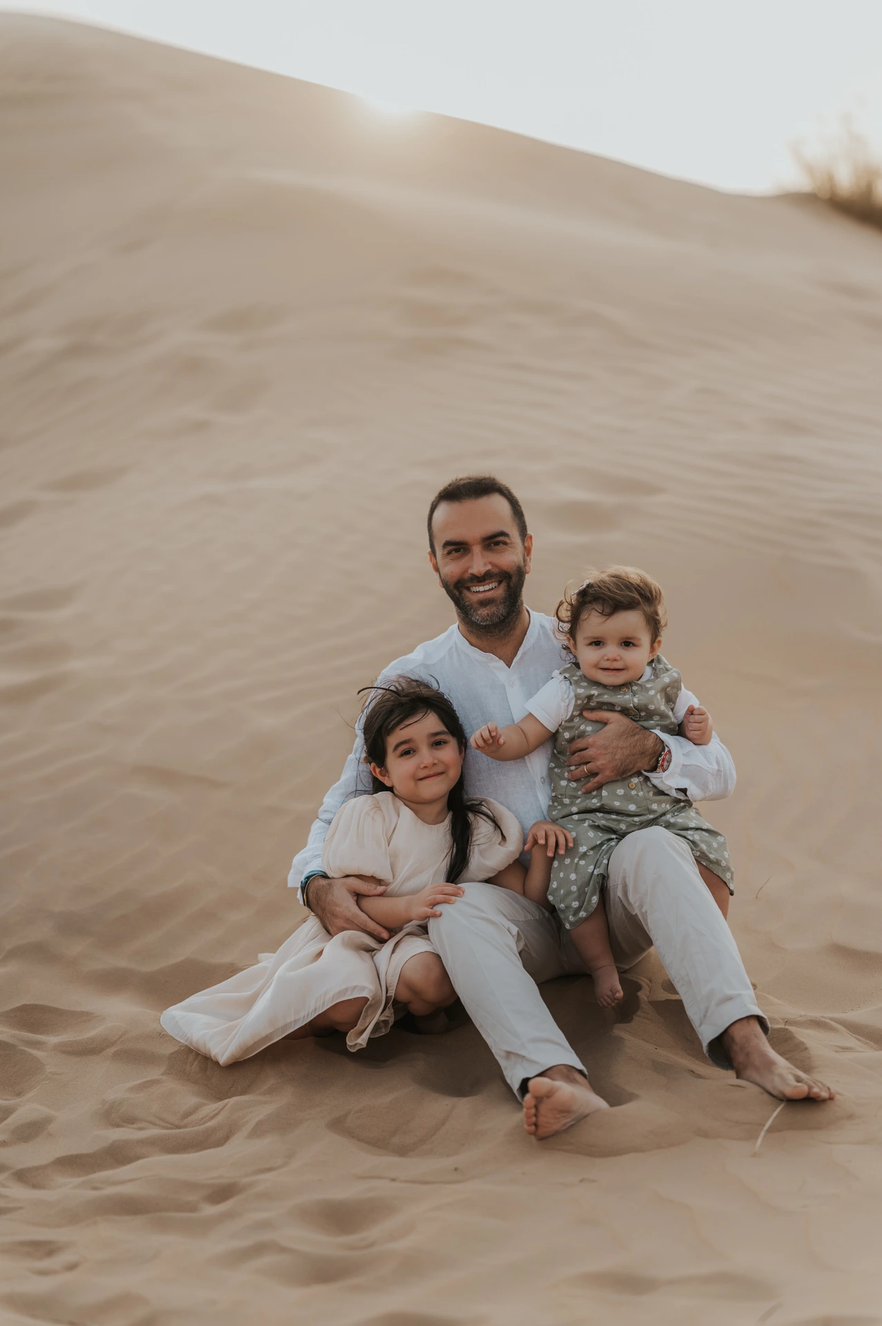 Smiling father and two young children sit on Dubai desert dunes during a warm, candid family photoshoot session.