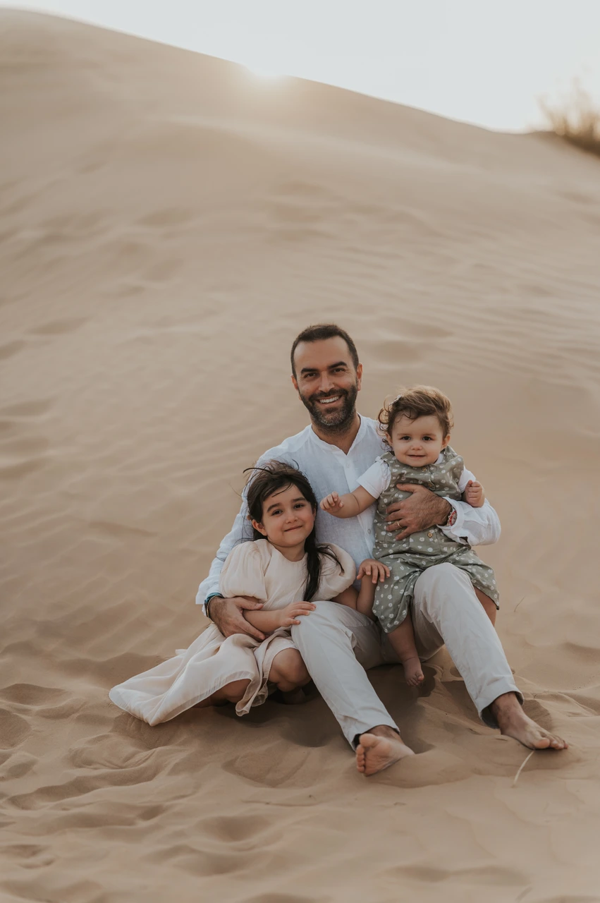 Smiling father and two young children sit on Dubai desert dunes during a warm, candid family photoshoot session. Smiling father and two young children sit on Dubai desert dunes during a warm, candid family photoshoot session.