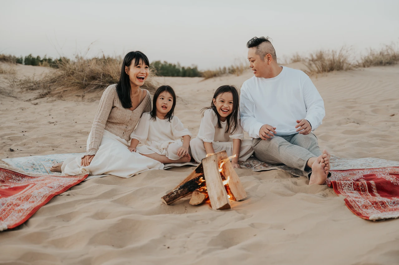 Family of four smiling beside a small campfire on desert blankets during a Dubai family photoshoot at sunset. Family of four smiling beside a small campfire on desert blankets during a Dubai family photoshoot at sunset.
