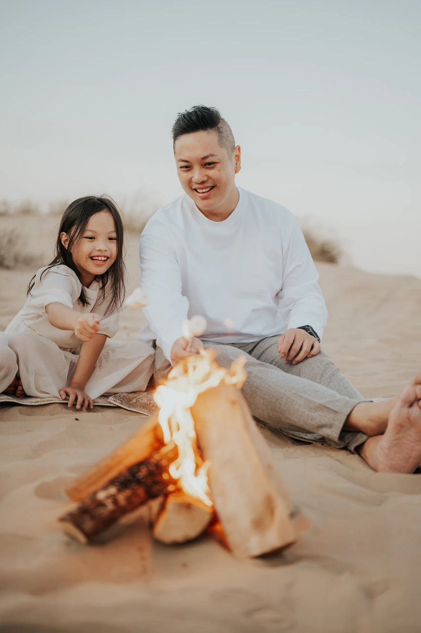 A father and daughter smile beside a campfire on Dubai desert sand during a relaxed family photo session. A father and daughter smile beside a campfire on Dubai desert sand during a relaxed family photo session.