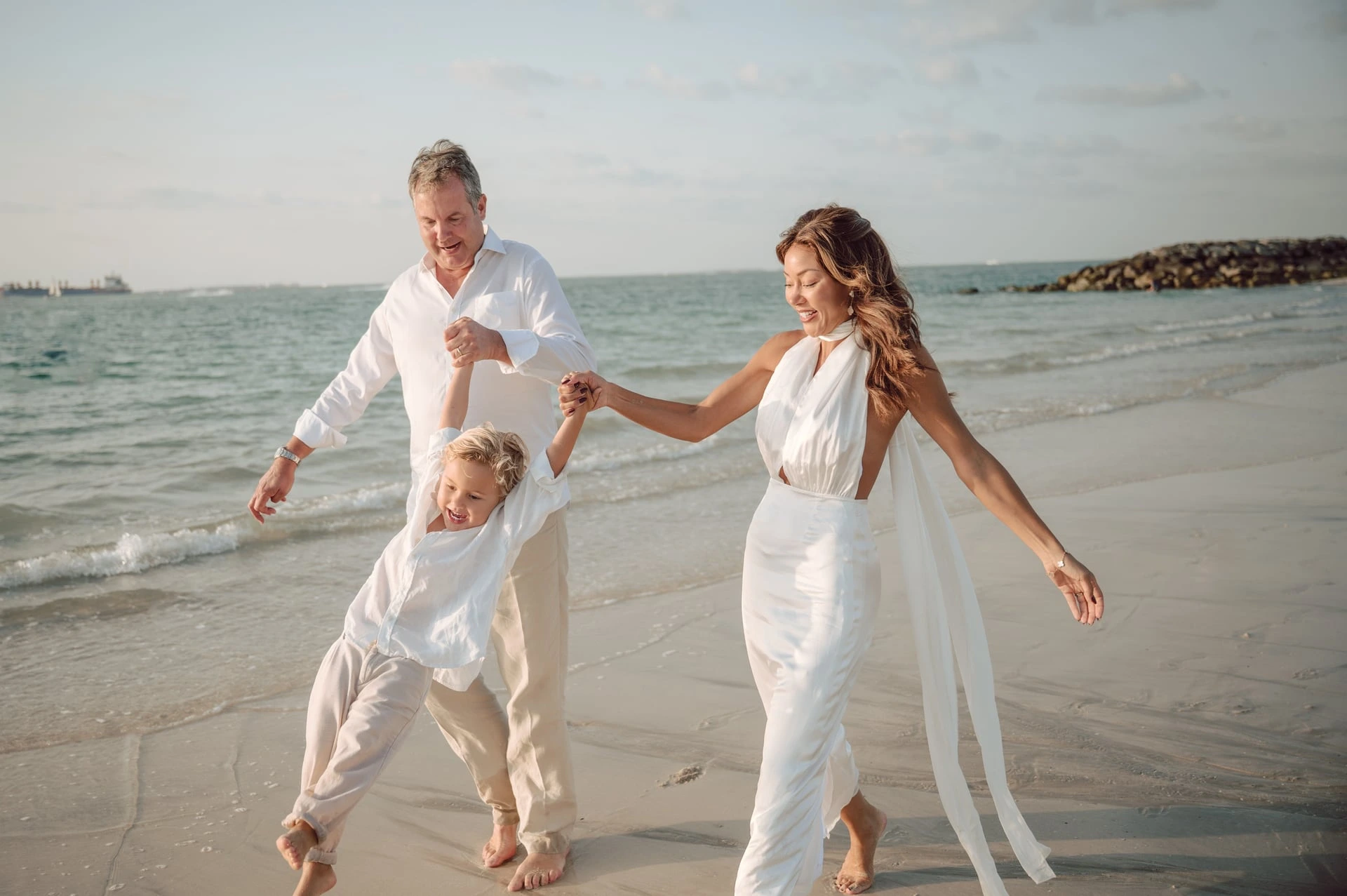 New Grandparent Gift Ideas to Celebrate Their Grandchild Parents walk along the beach holding their young child’s hands.