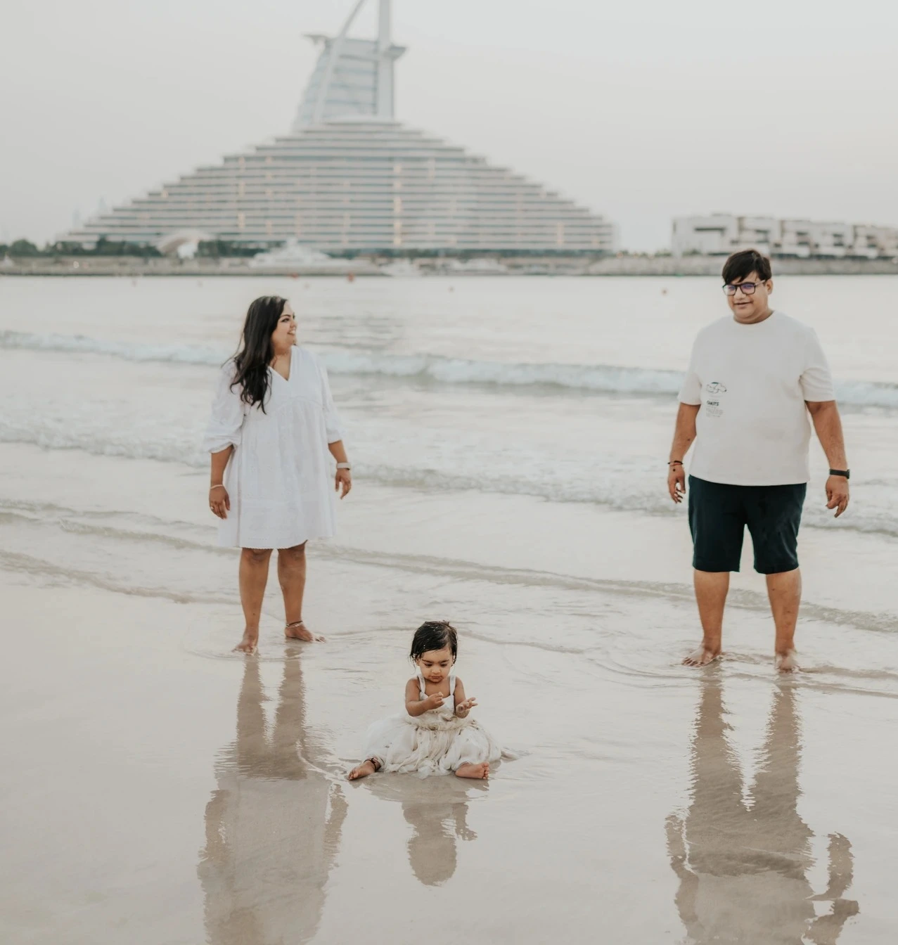 Parents stand at the shoreline while their toddler plays in shallow water on Dubai beach during a natural family photoshoot session.