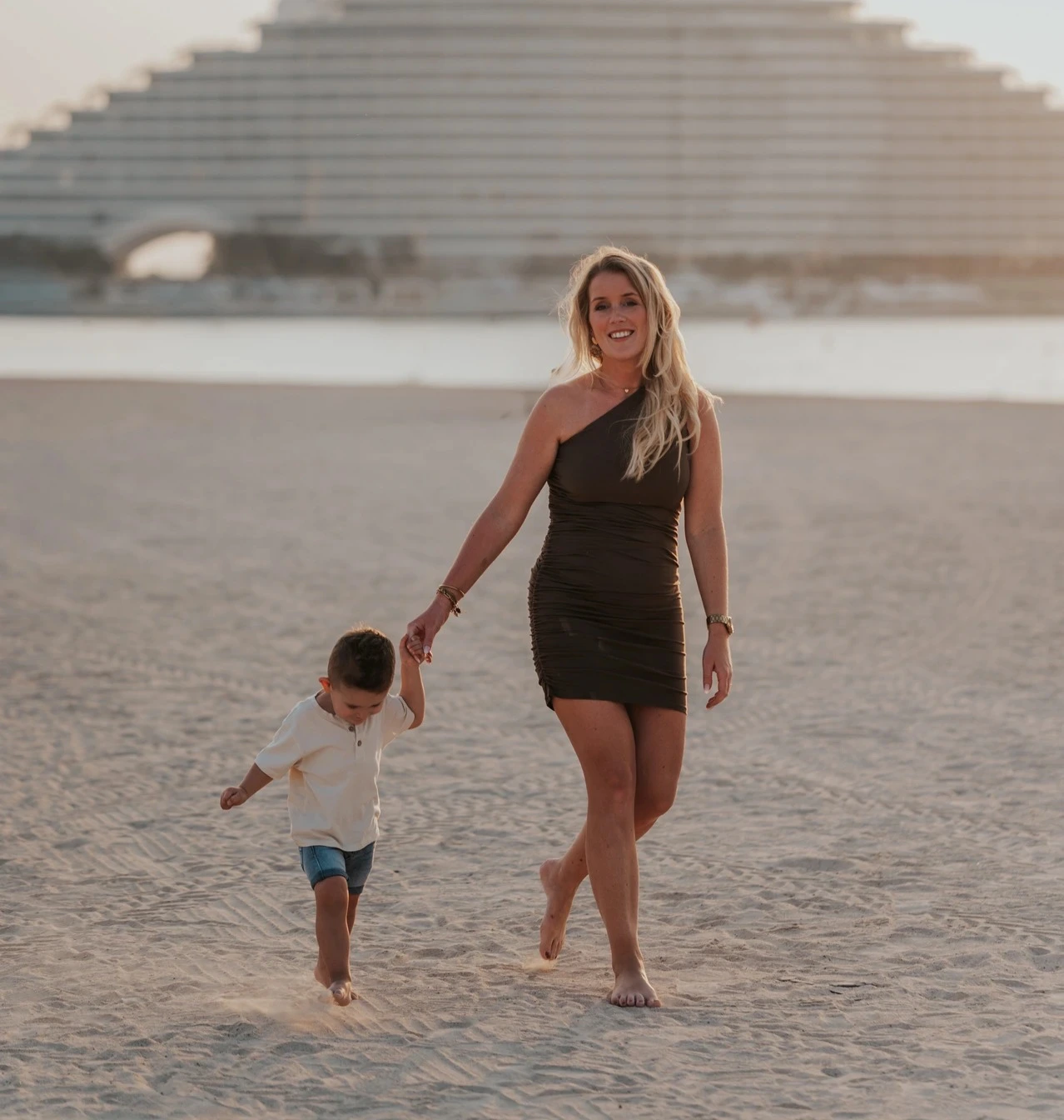 A mother holds her toddler’s hand while walking on Dubai beach with Jumeirah Beach Hotel behind them during a family photoshoot.