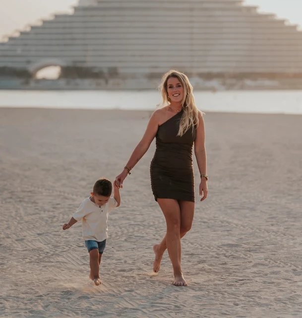 A mother holds her toddler’s hand while walking on Dubai beach with Jumeirah Beach Hotel behind them during a family photoshoot.