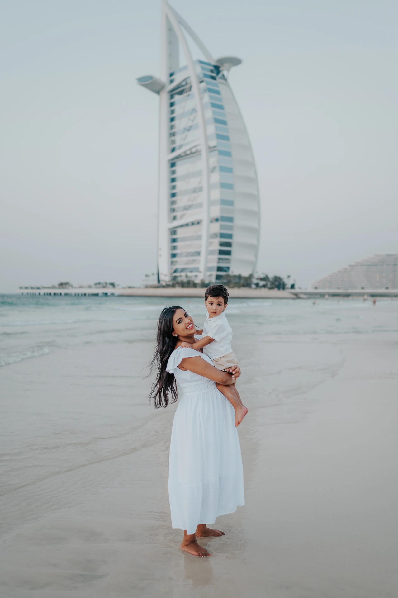 Family photoshoot in Dubai - Burj Al Arab toddler portrait A mother holds her toddler on Dubai beach with Burj Al Arab in the background during a family photoshoot.