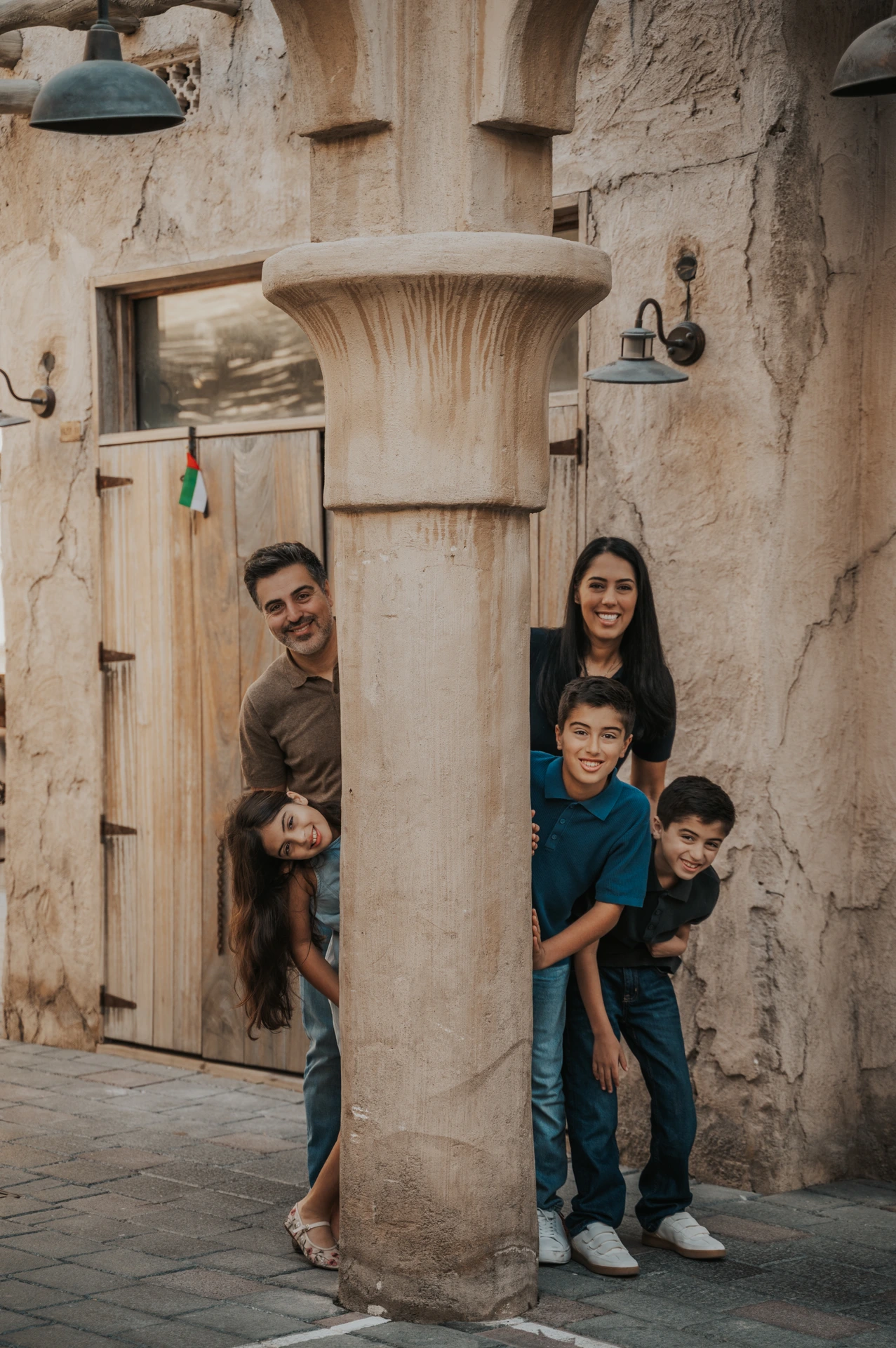 Family of five peeking around a stone column in rustic alleyway during playful Dubai family photoshoot session.