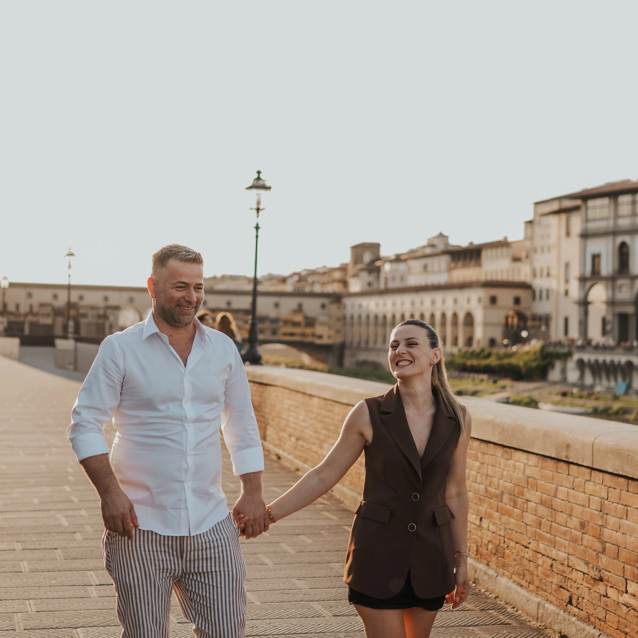 Una coppia sorridente cammina mano nella mano lungo l’Arno, con Ponte Vecchio sullo sfondo, durante un servizio fotografico a Firenze.