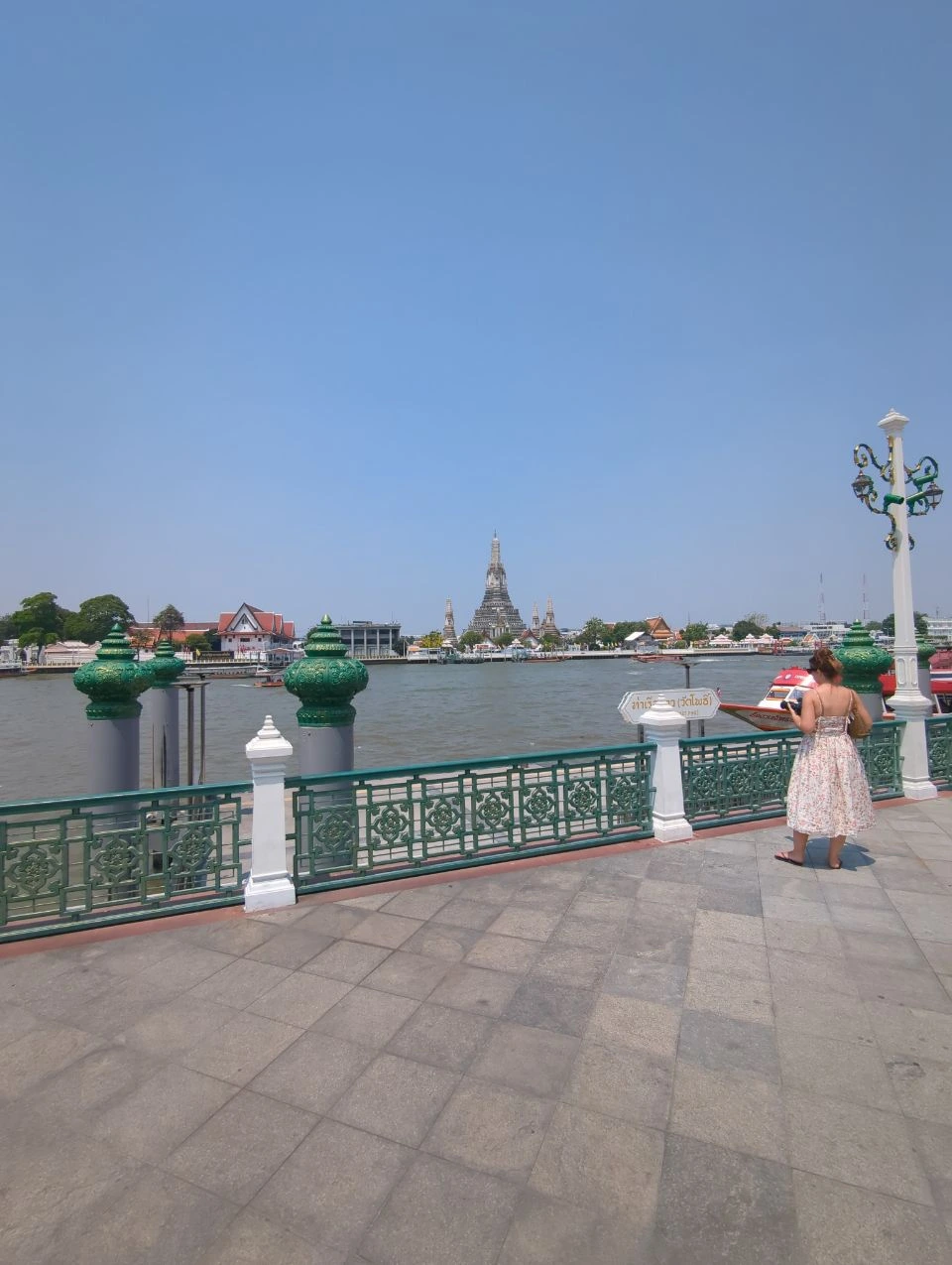 Wat Pho vs Wat Arun Photos - Riverside Platform A visitor stands on a Bangkok riverside platform with Wat Arun visible across the Chao Phraya River.