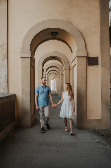 The couple walk hand in hand beneath a long series of arches during their Florence couple photoshoot.