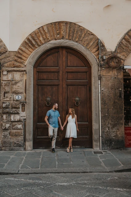 Florence couple photoshoot beneath an arched wooden doorway The couple hold hands beneath a large arched wooden doorway in a stone wall during their Florence photoshoot.