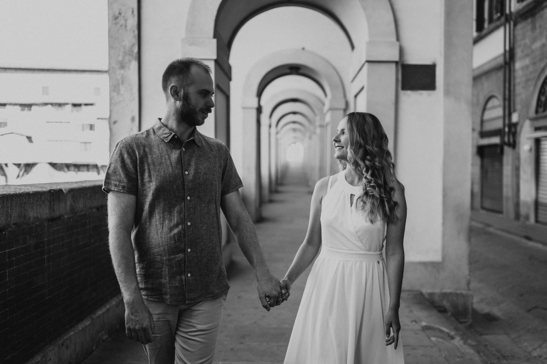 Black-and-white photograph of the couple walking through an arched passage during their Florence couple photoshoot.