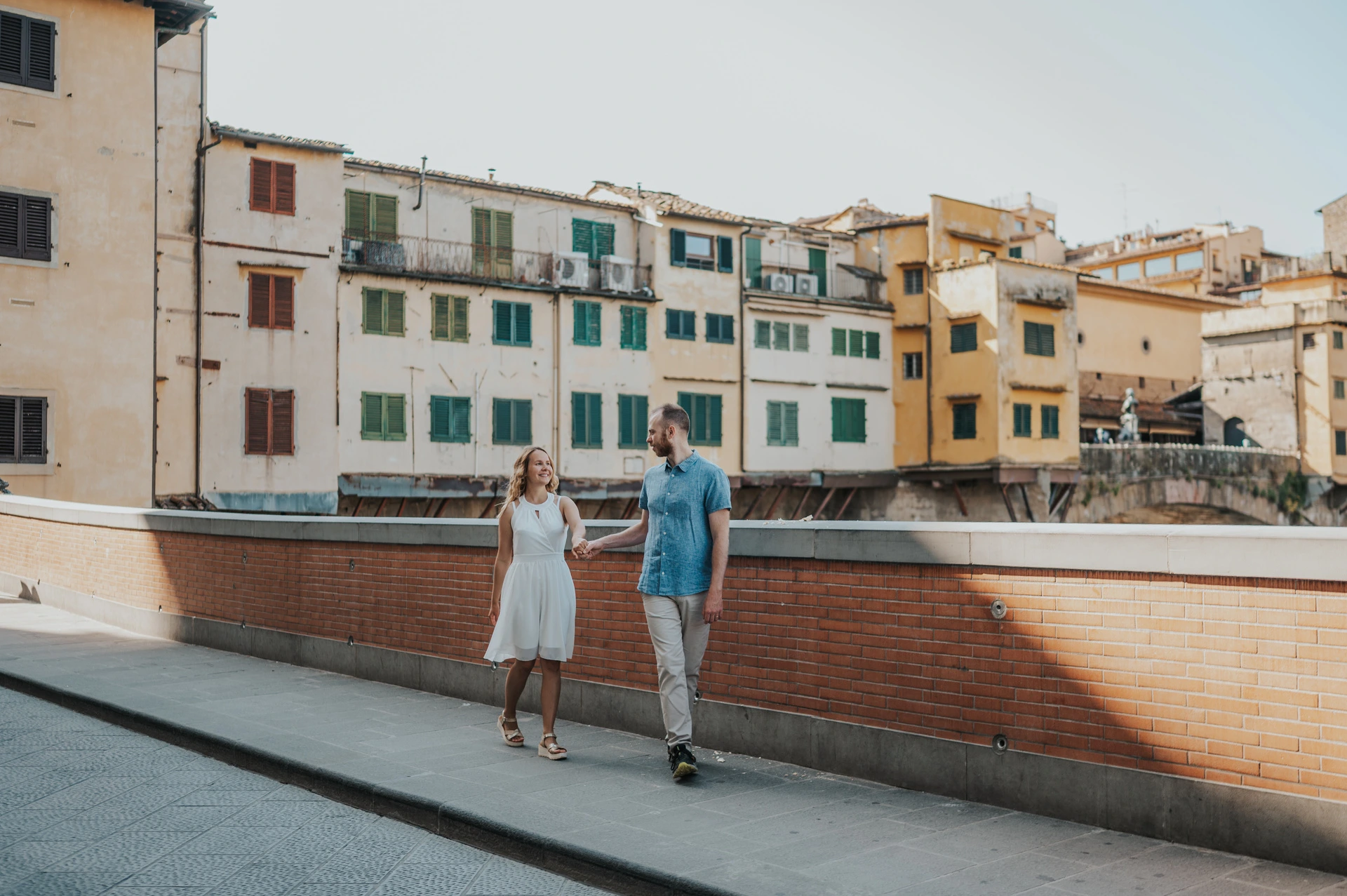 The couple hold hands by a brick parapet, with colourful buildings across the Arno, during their Florence photoshoot.