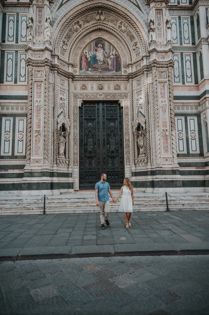 A wide Duomo doorway portrait from a Florence couple photoshoot The couple walk hand in hand below the Duomo’s large carved doorway and patterned marble during their Florence photoshoot.