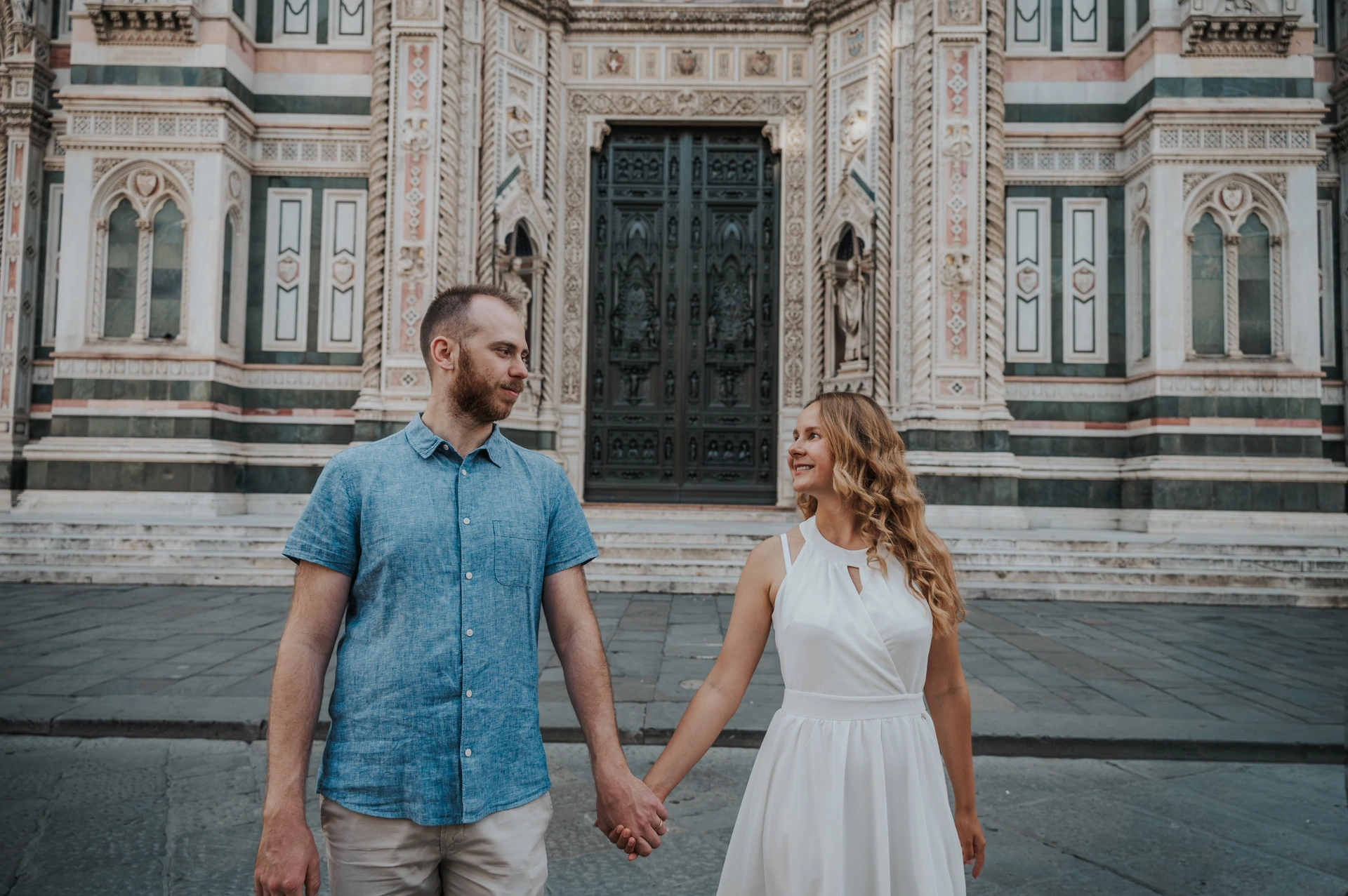 The couple hold hands and look towards each other, with the Duomo’s marble facade behind them, during their Florence photoshoot.