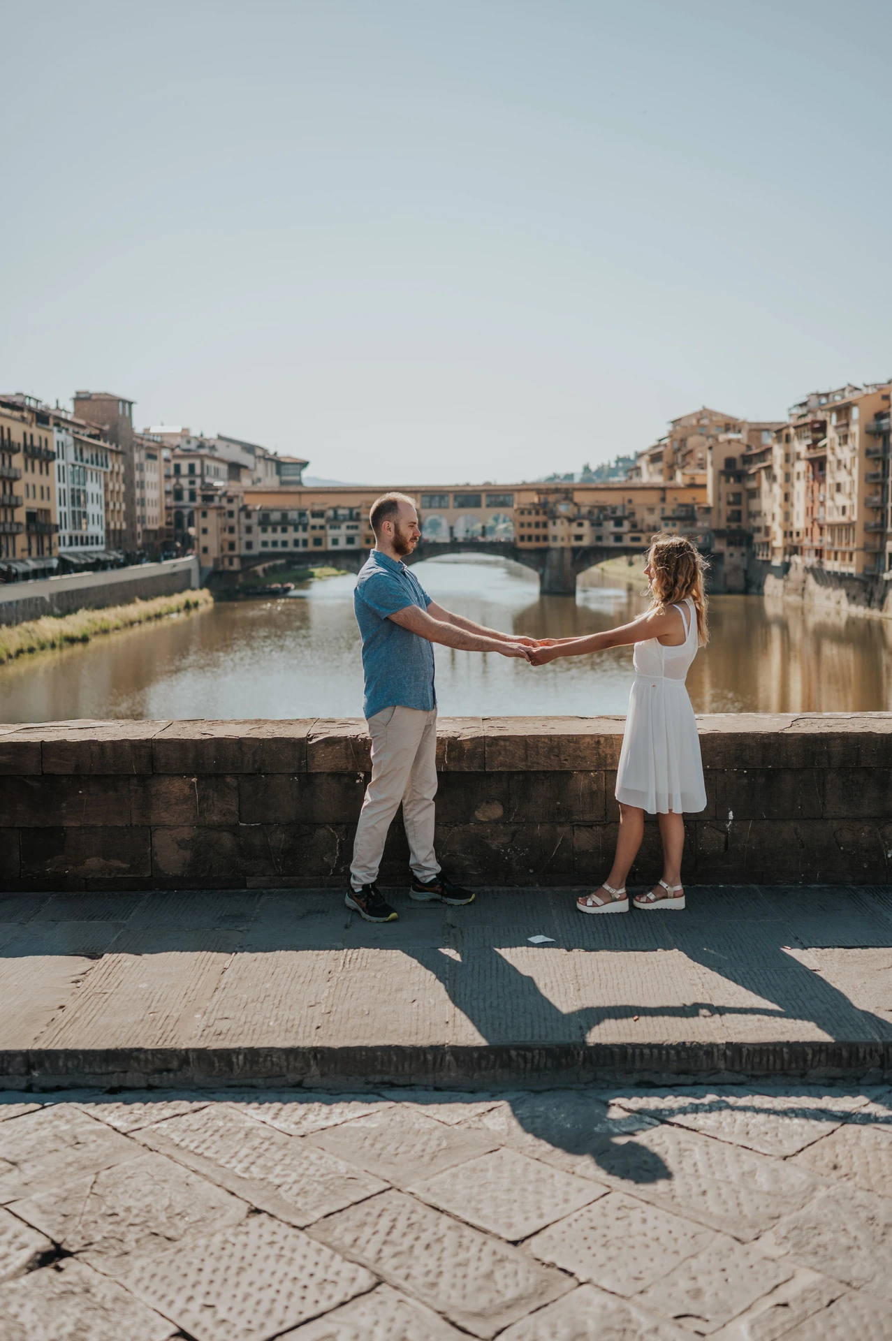 The couple face each other and hold hands by the river parapet, with Ponte Vecchio behind them, during their Florence photoshoot.