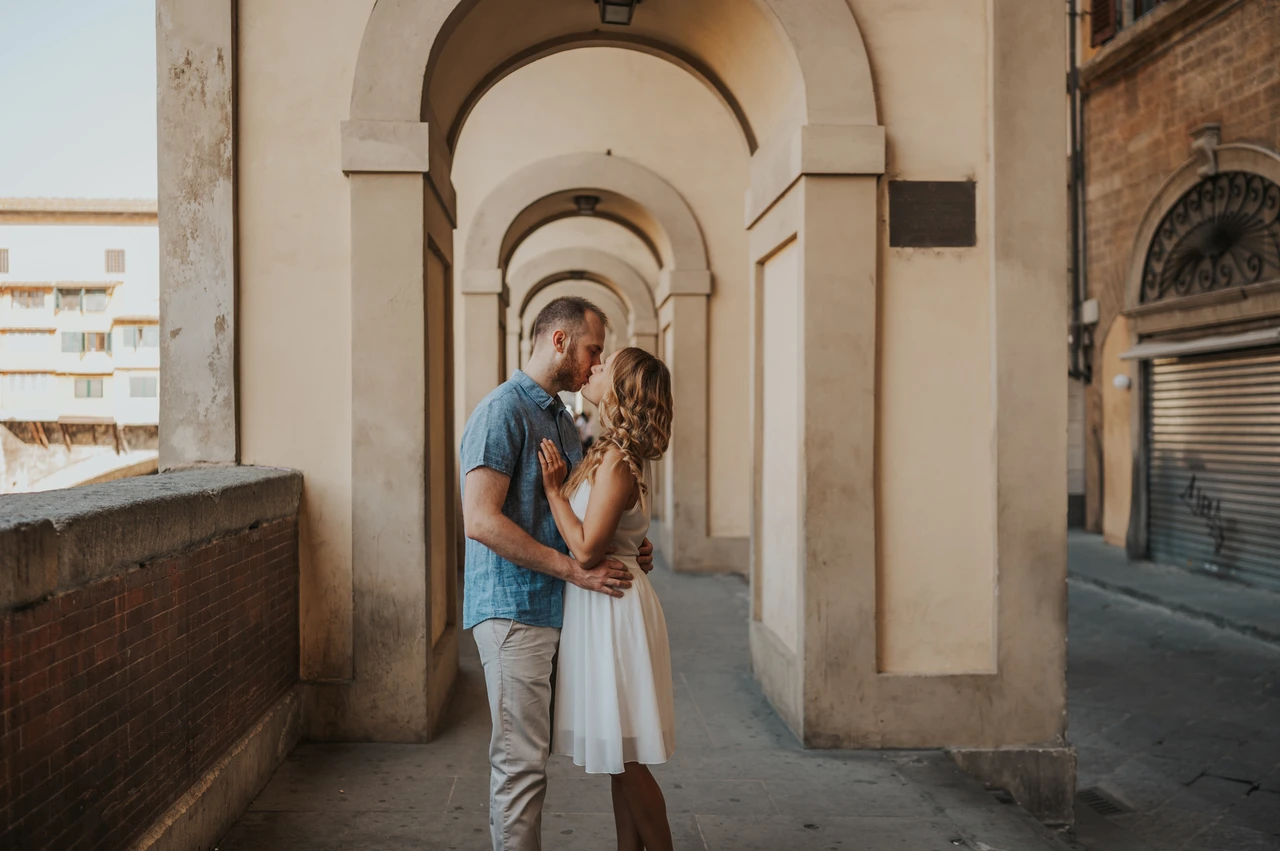 A kiss beneath the arches during a Florence couple photoshoot The couple kiss beneath a series of pale arches beside a brick parapet during their Florence photoshoot.