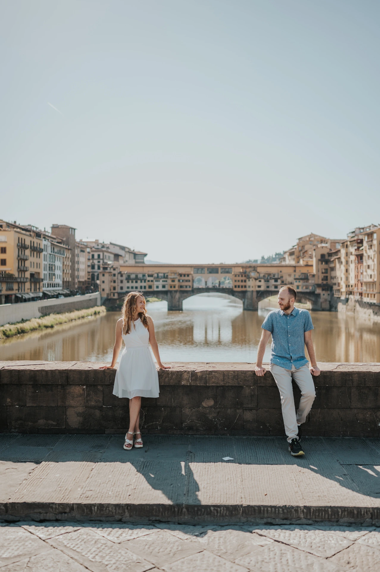 The couple pause beside the river parapet with Ponte Vecchio and the Arno behind them during their Florence photoshoot.