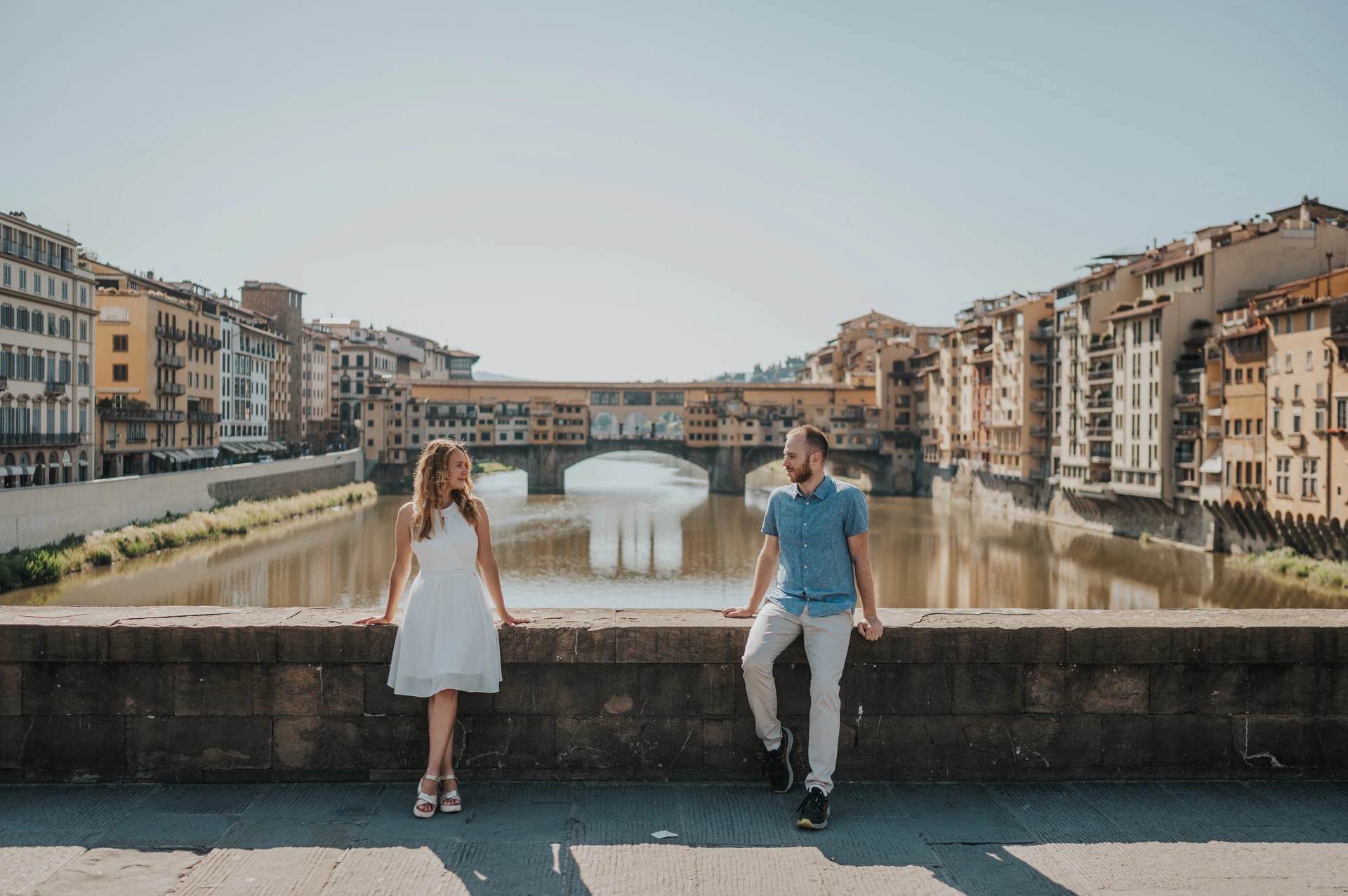 The couple stand by the stone parapet above the Arno, with Ponte Vecchio in the background, during their Florence couple photoshoot.